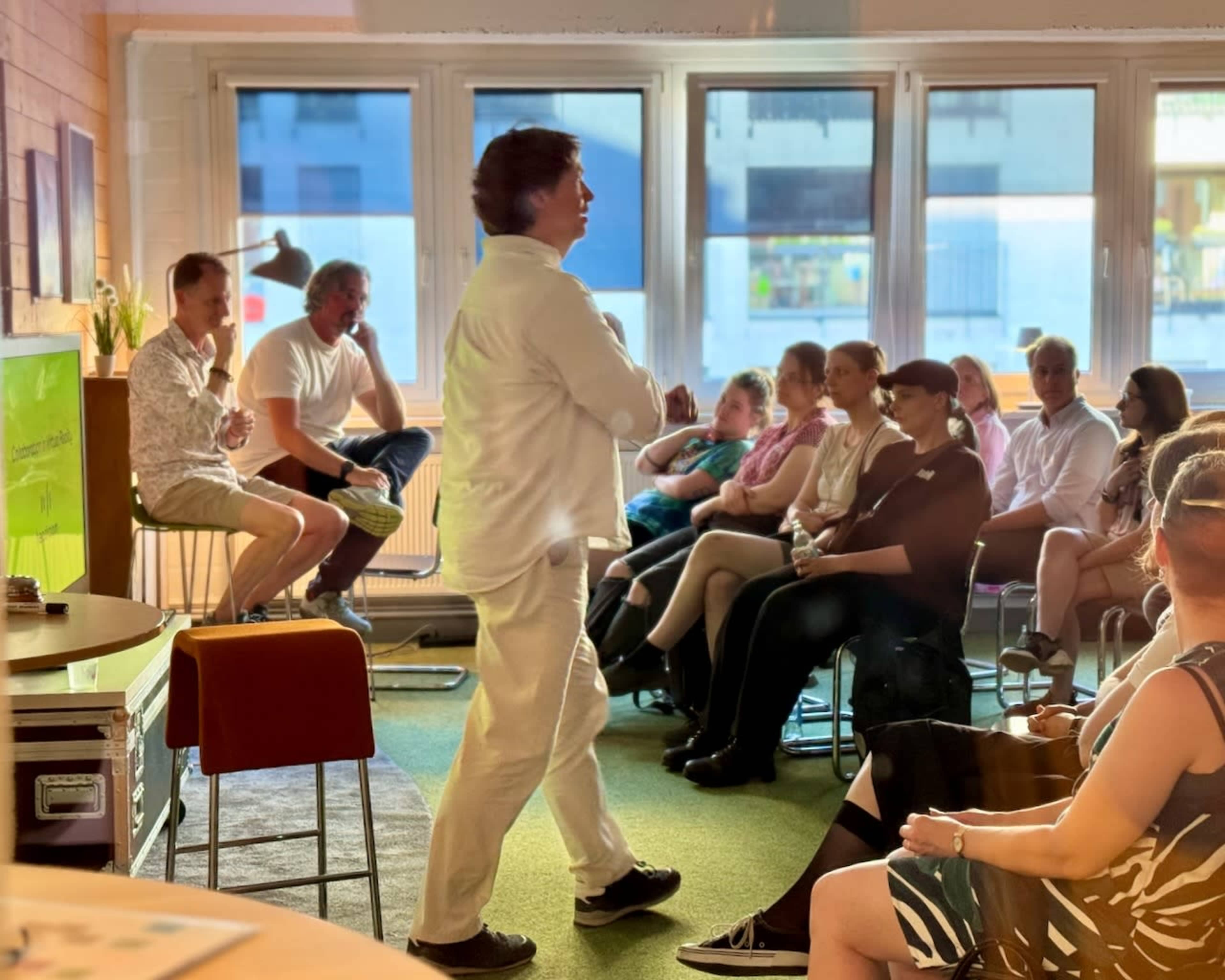 A speaker in white attire presents to an audience seated on chairs in a brightly lit room.