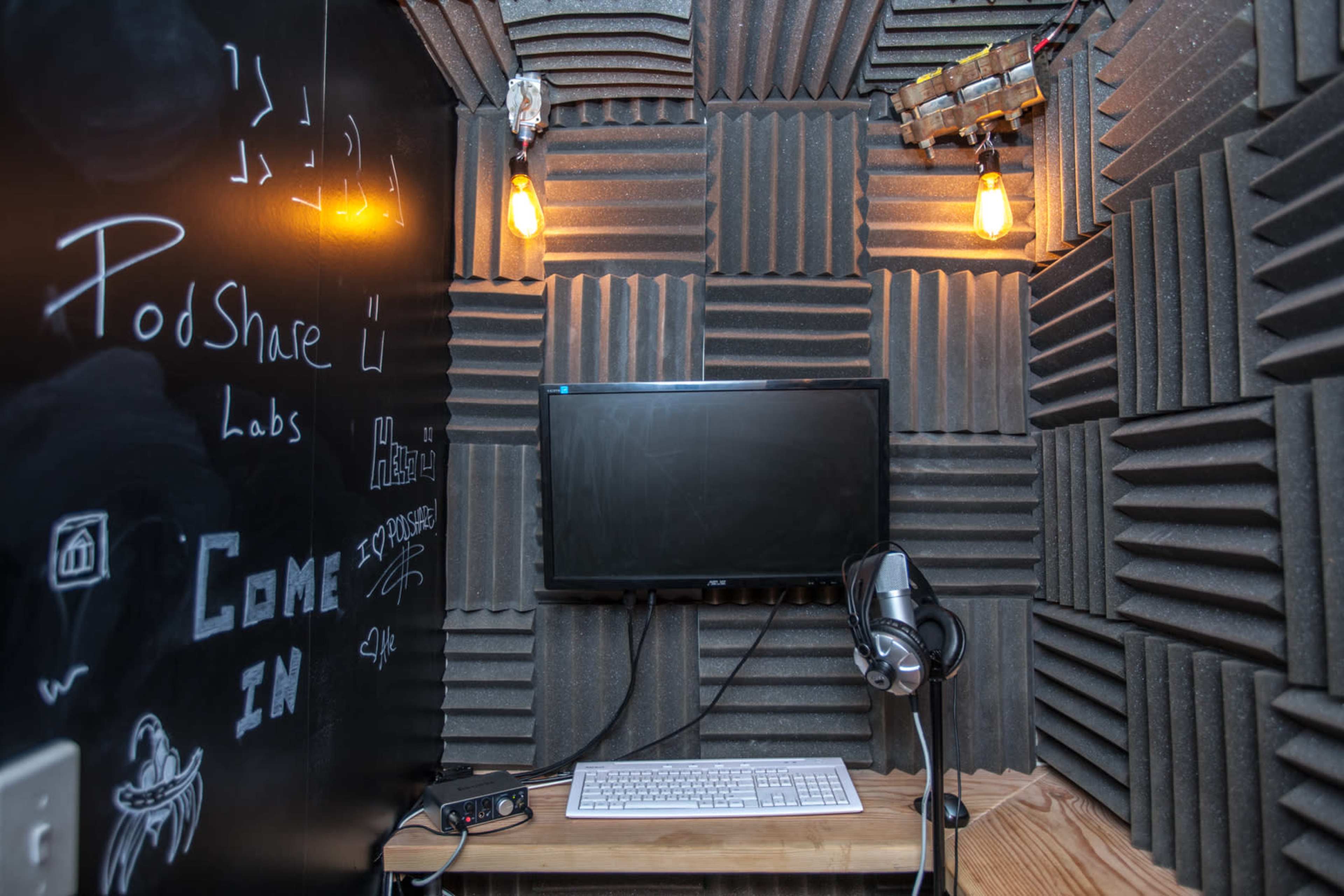 A small recording studio space with soundproofing foam on the walls, a monitor on a wooden desk, a microphone, and two exposed light bulbs hanging from the ceiling.