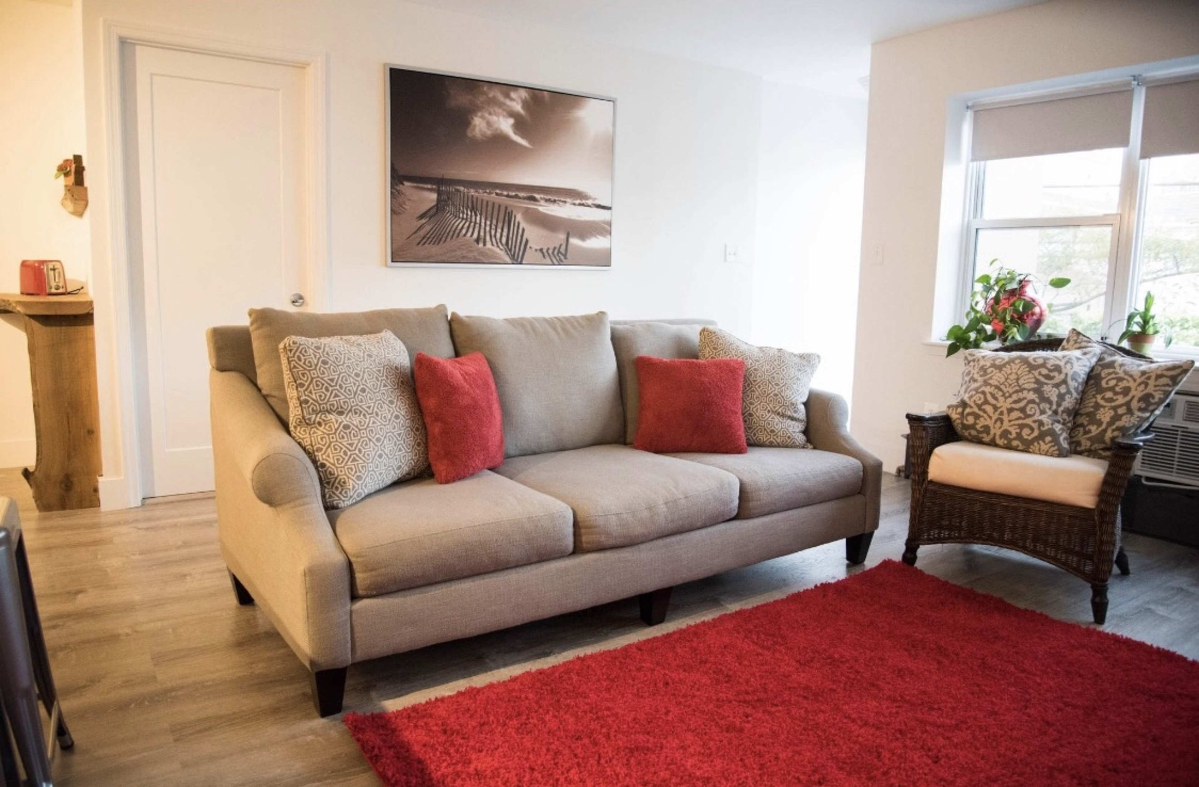 A living room with a light beige sofa adorned with four decorative pillows, a red area rug on the floor, and a wall featuring a black-and-white photograph.