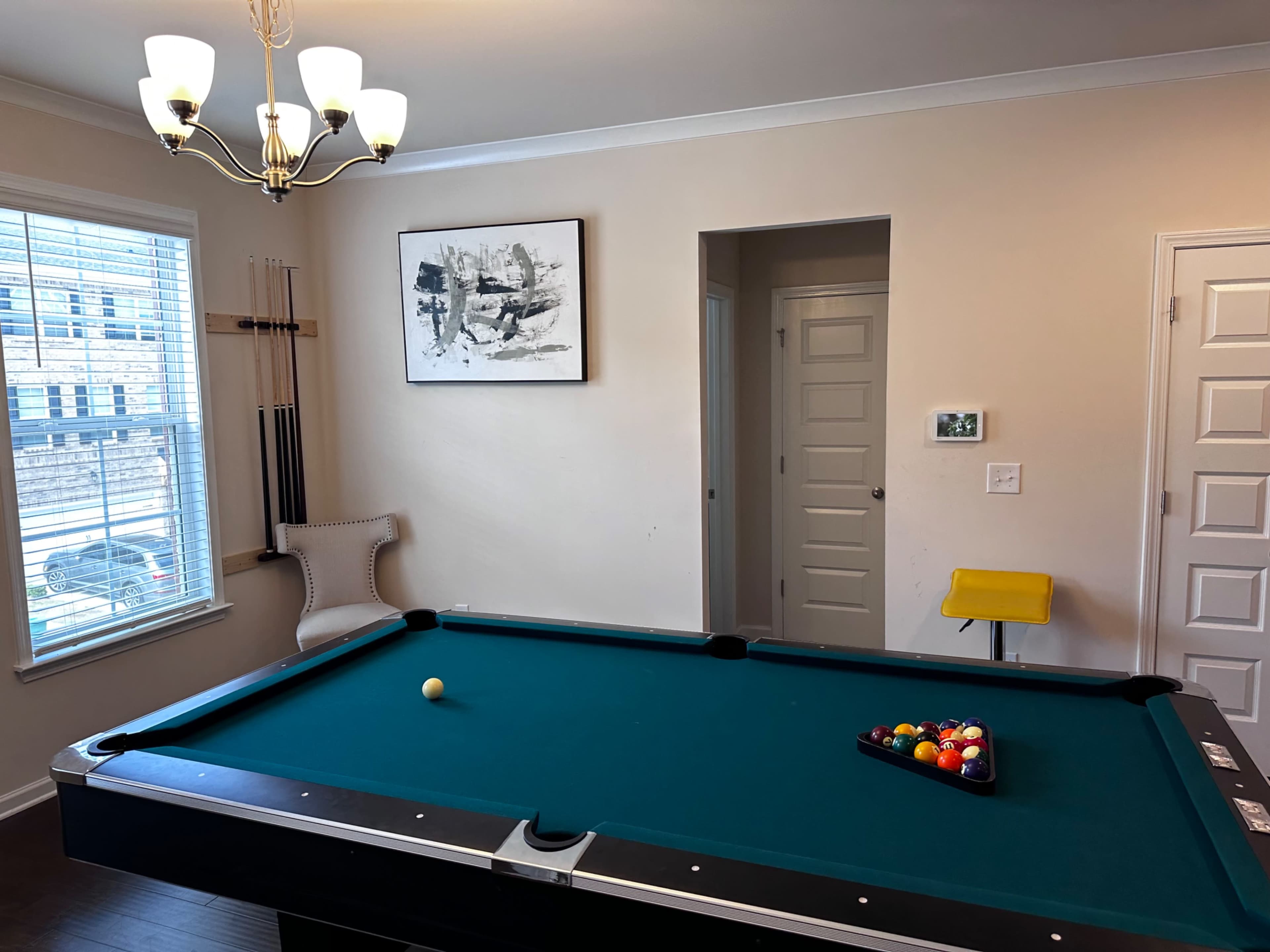 A well-lit room featuring a billiard table, a set of pool cues on the wall, and a small yellow stool near a doorway.