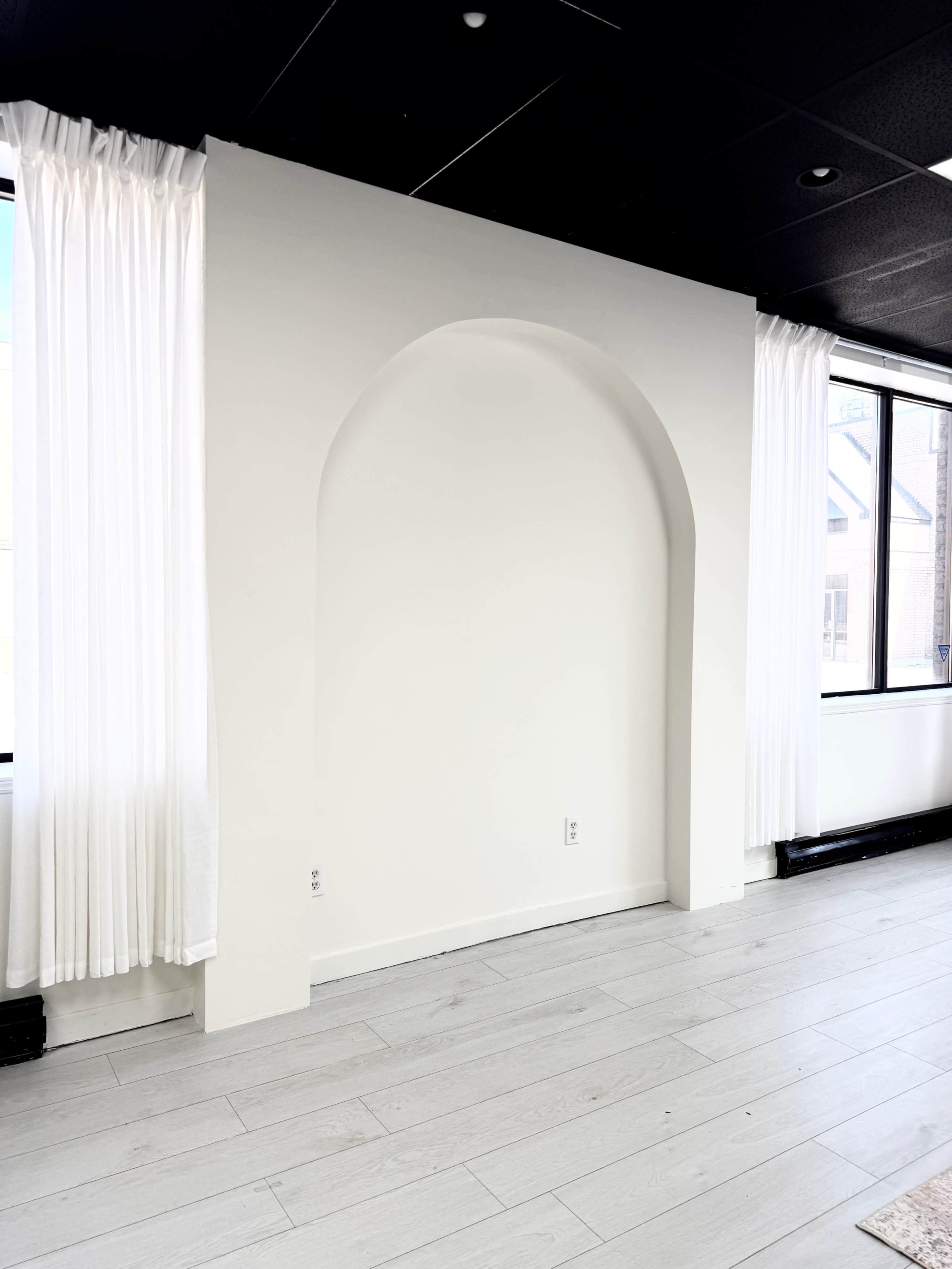The image shows a blank white wall with an arched recess and large windows covered with sheer white curtains.