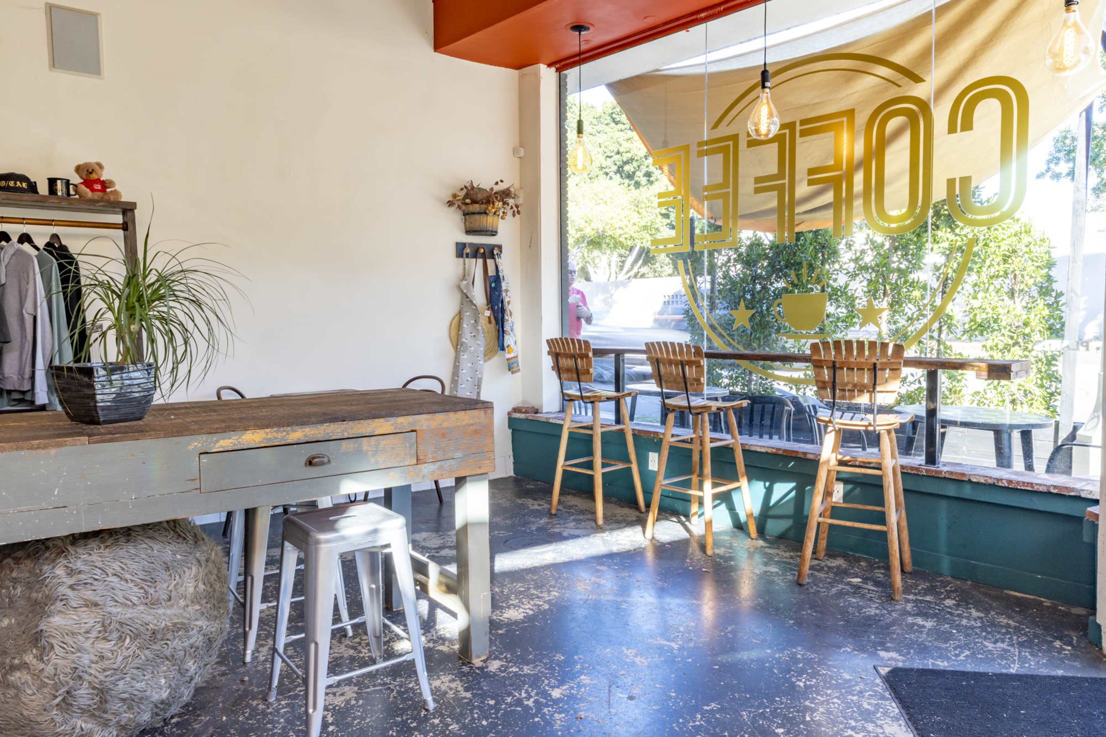 A coffee shop interior with a wooden table, high stools by a large window, and potted plants.