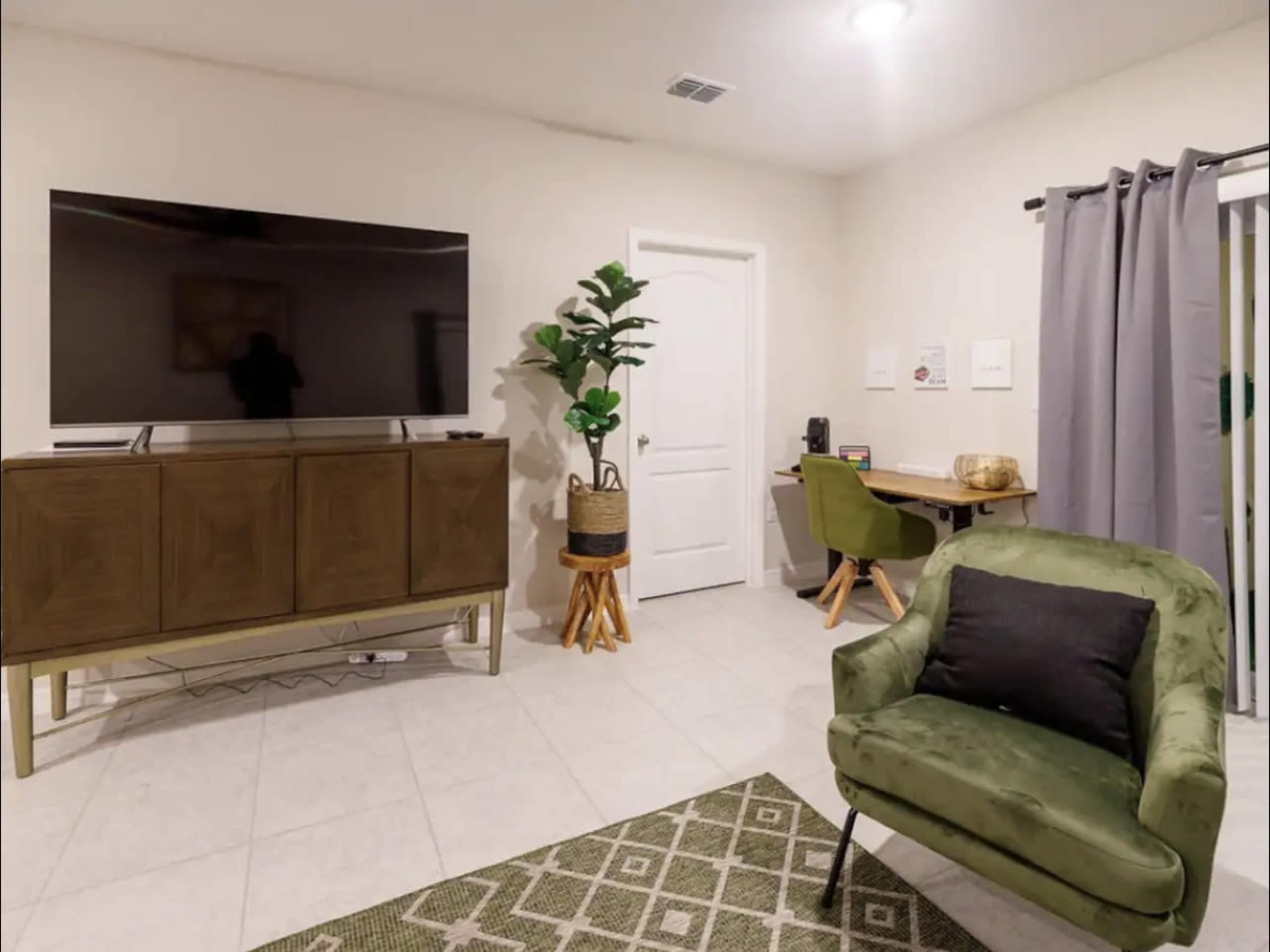 A room with a large TV mounted on the wall, a green armchair, a wooden cabinet, and a workspace featuring a desk and chair.