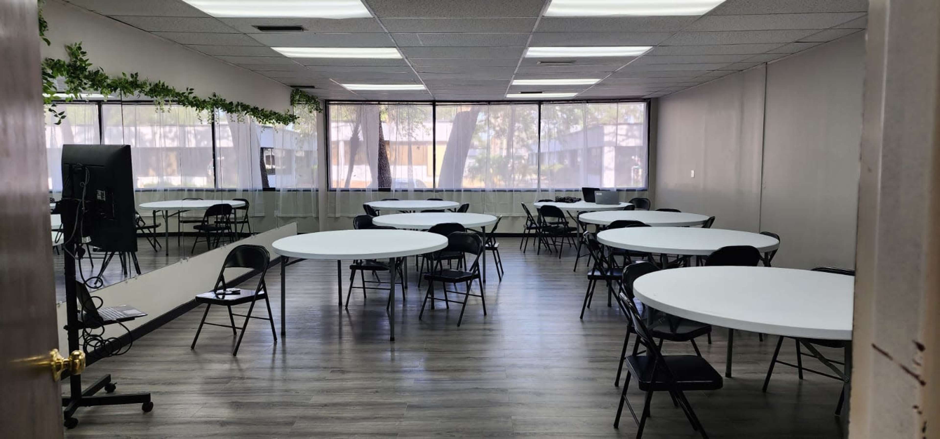 The image shows a spacious meeting room with several round tables and black chairs arranged throughout, illuminated by large windows.