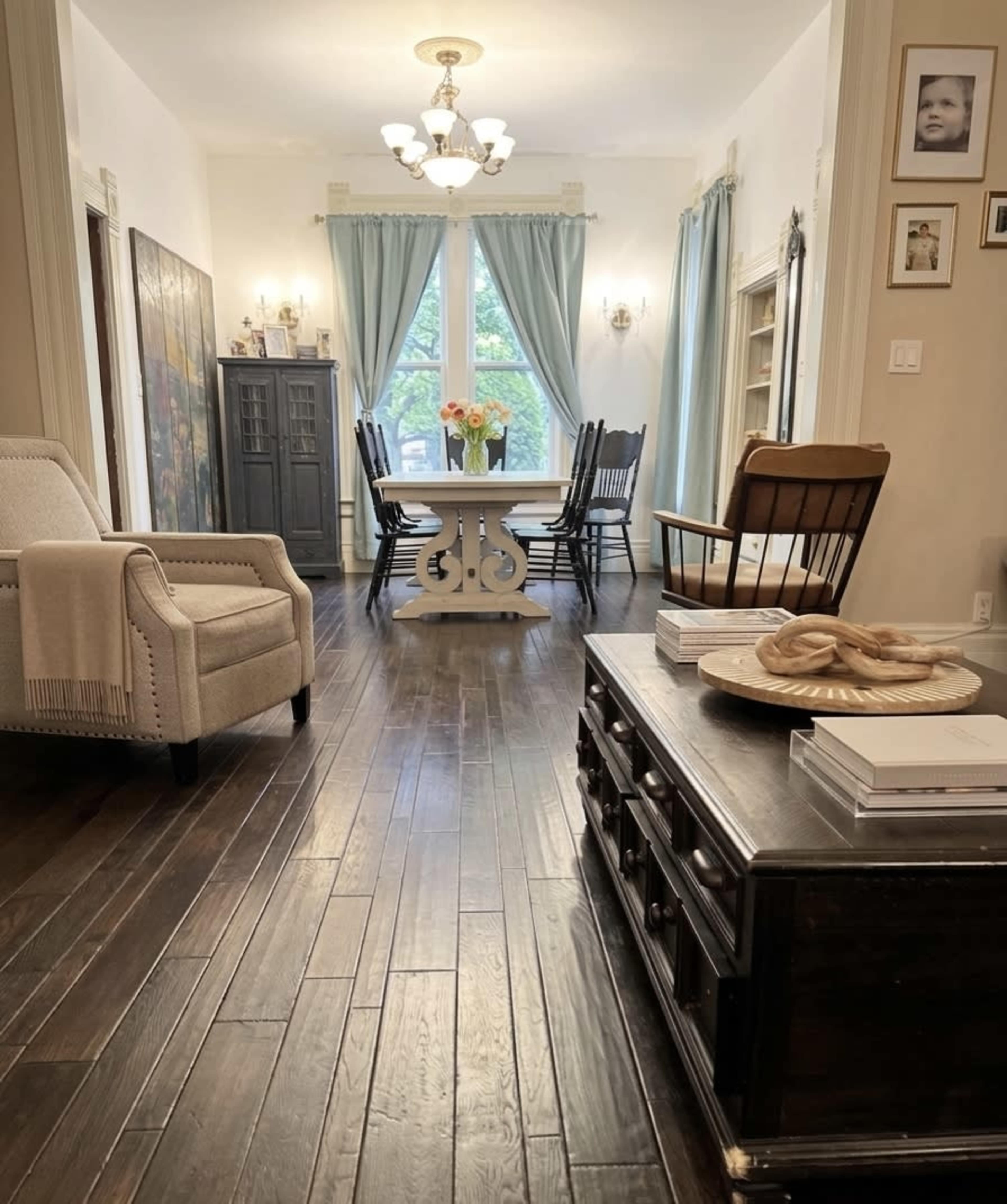 The image shows a living space with wooden floors, a central dining table surrounded by chairs, and large windows dressed with light blue curtains.
