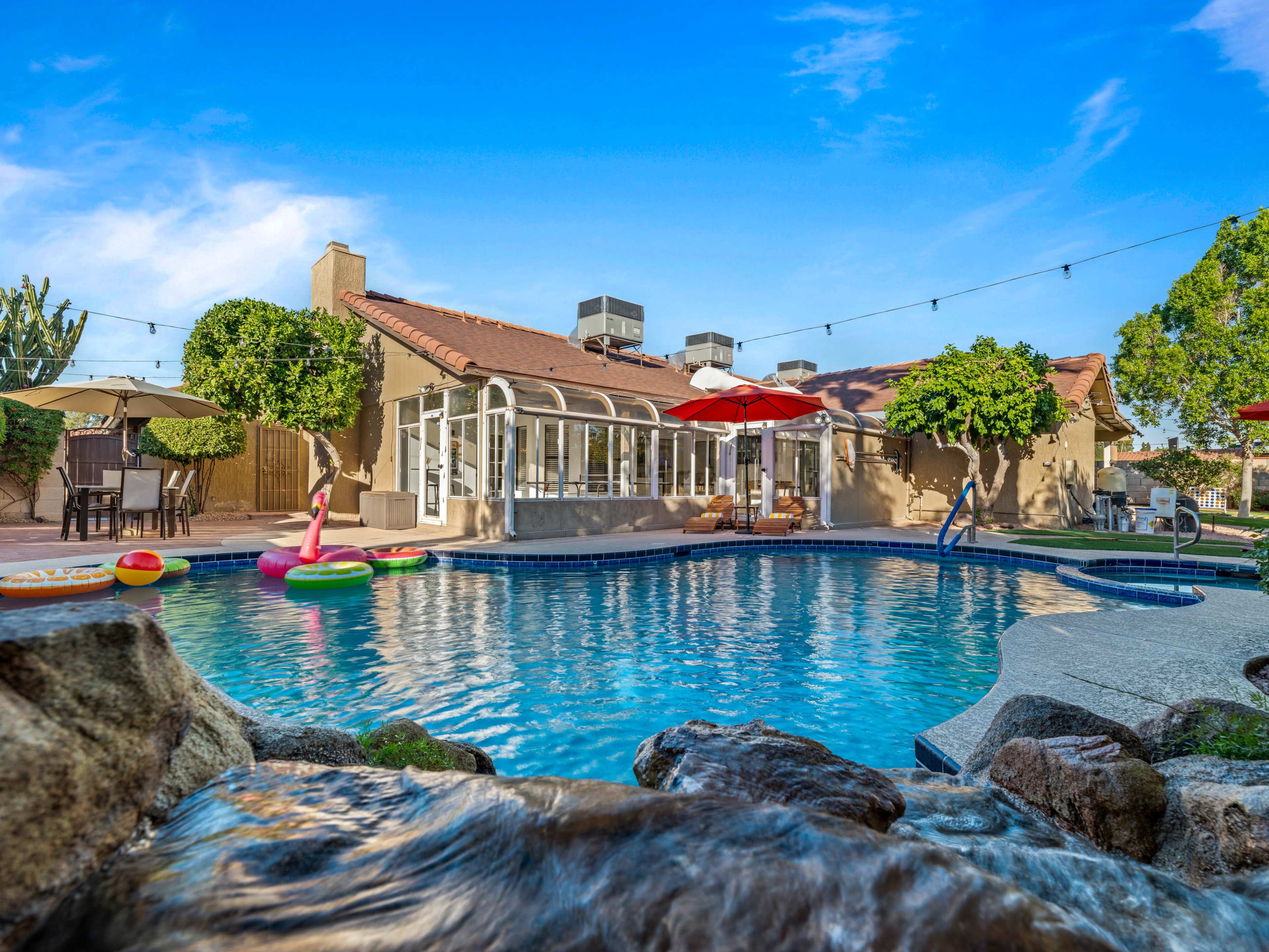 A clear pool with inflatable floats is situated in a backyard alongside a house with a patio and umbrellas.
