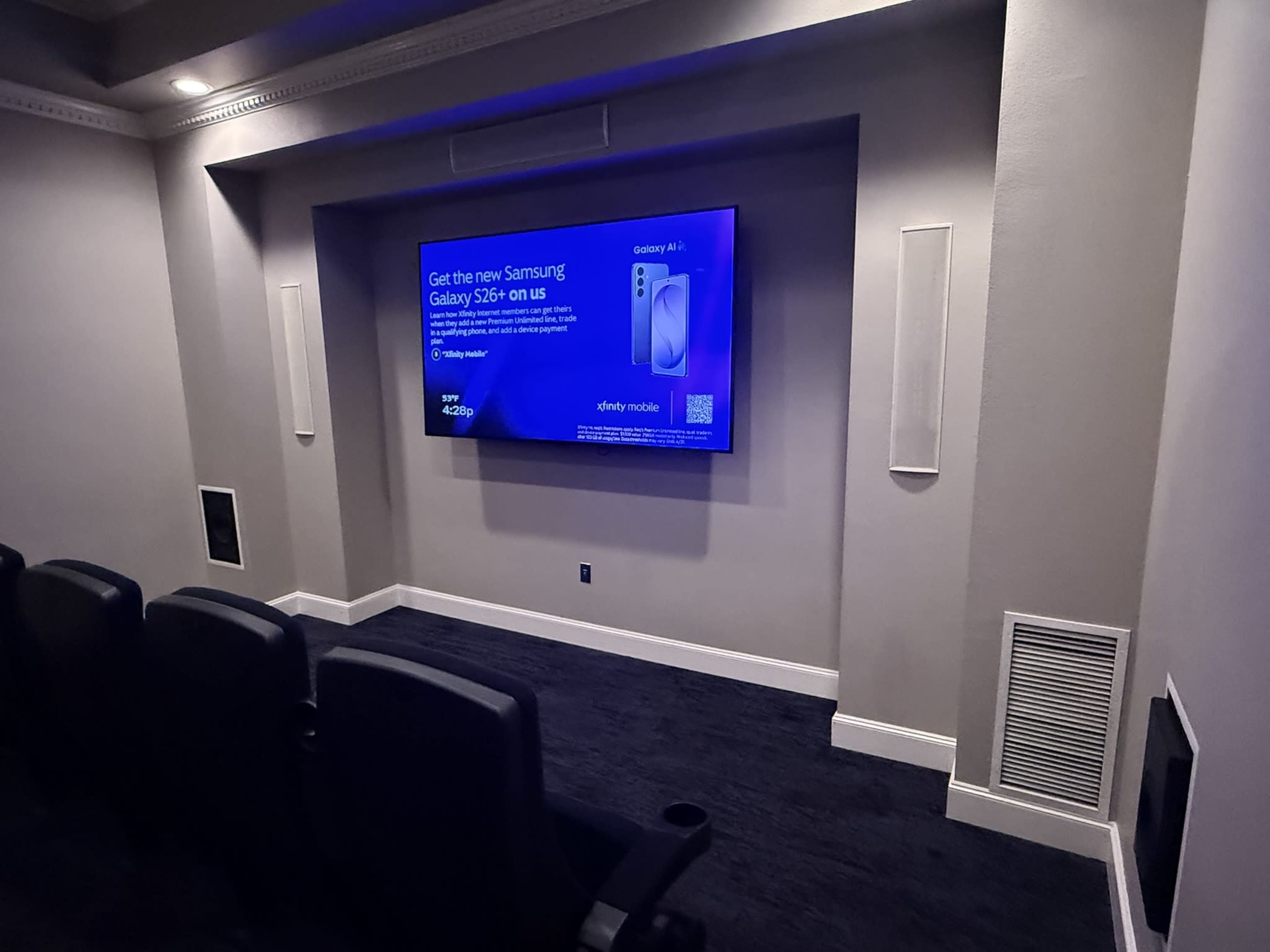 The image shows a home theater room with a large screen displaying an advertisement for the Samsung Galaxy S24 on a wall, and black seating in front.