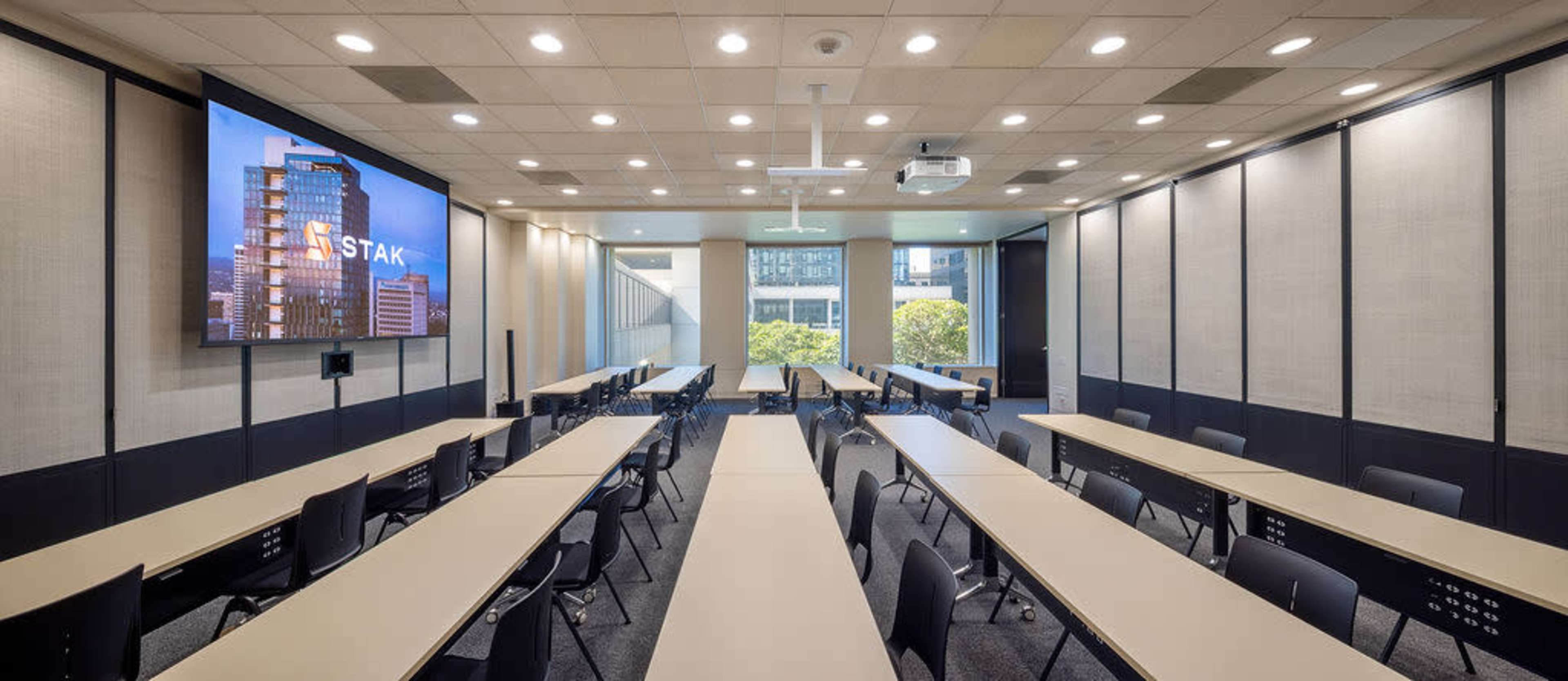 The image shows a modern conference room with multiple long tables arranged in a U-shape, a large screen displaying a building, and large windows allowing natural light.