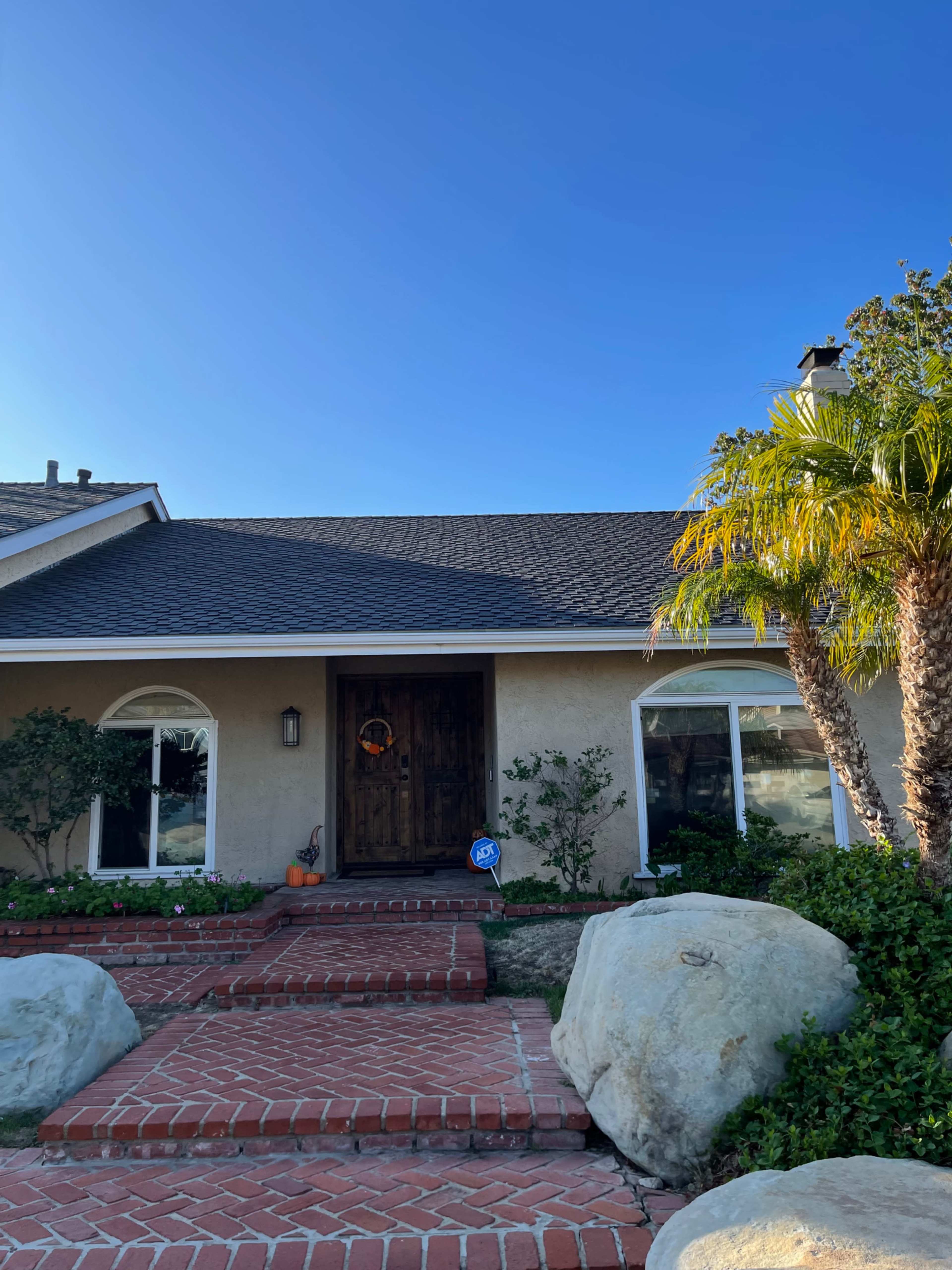 A house with a wooden front door, surrounded by a landscaped garden featuring palm trees and large rocks, under a clear blue sky.