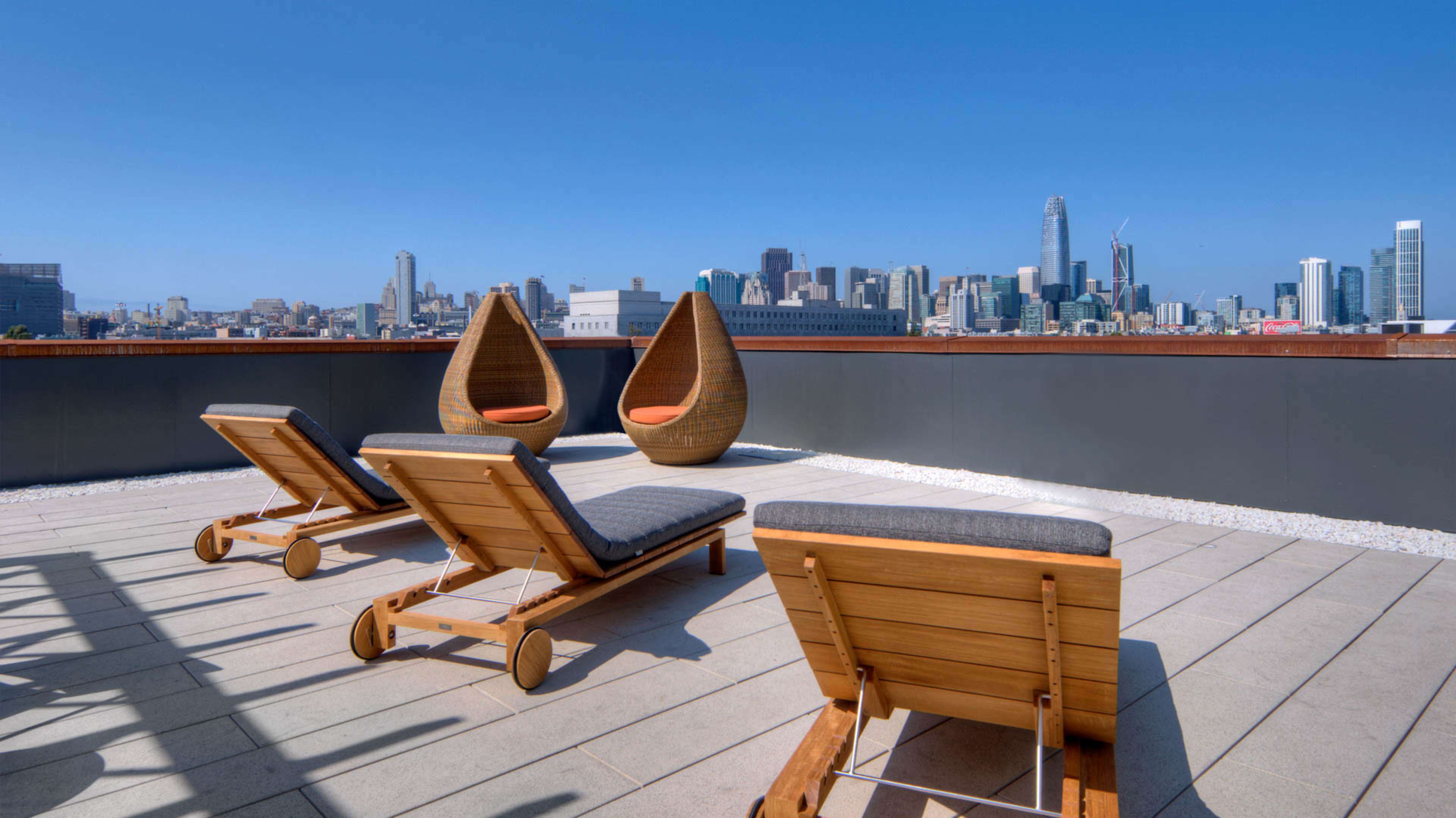 A rooftop terrace with two hanging chairs and three lounge chairs overlooking a city skyline on a clear day.