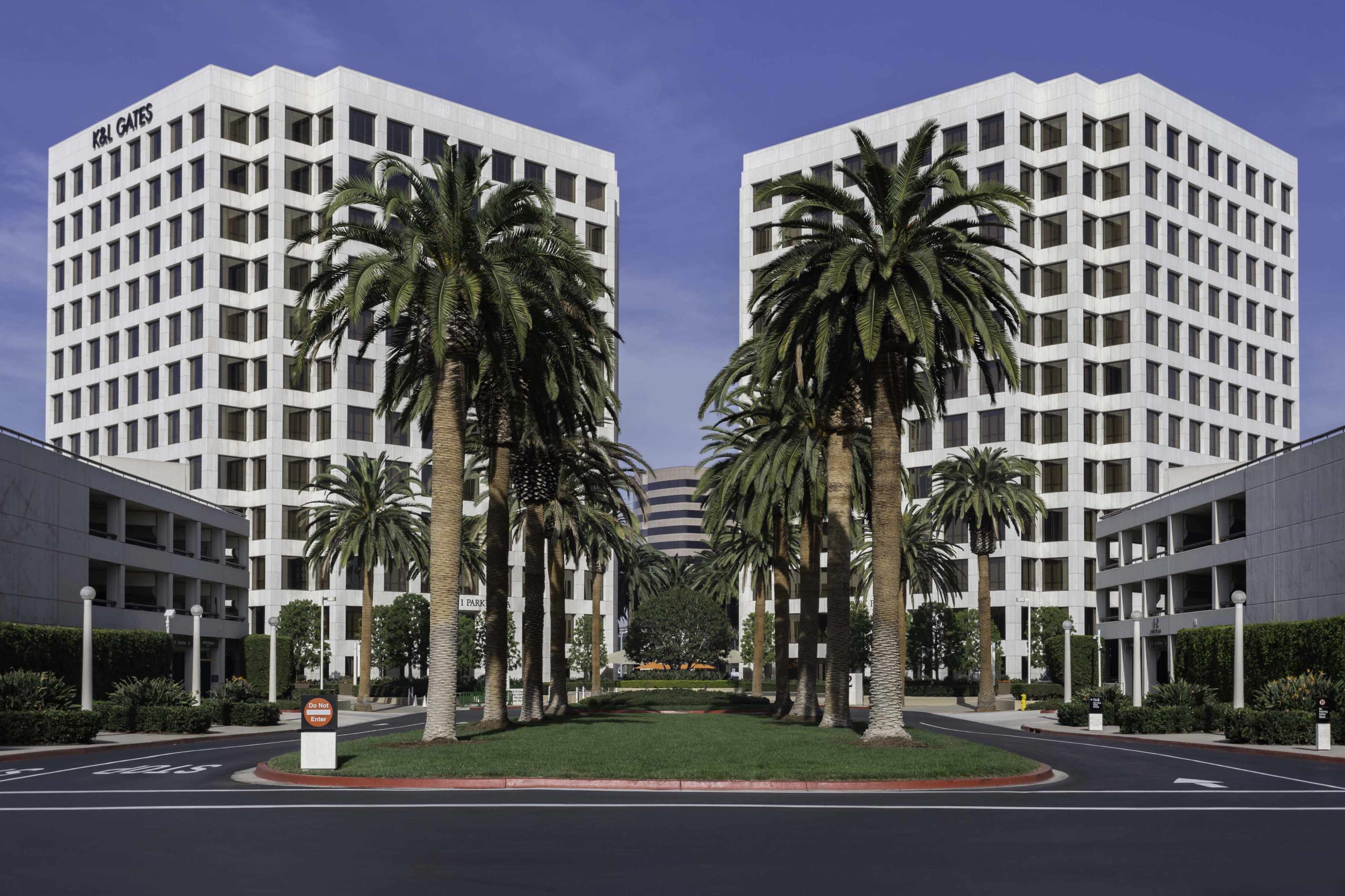 The image shows two identical office buildings flanked by palm trees, with a landscaped pathway in between leading to the entrance.