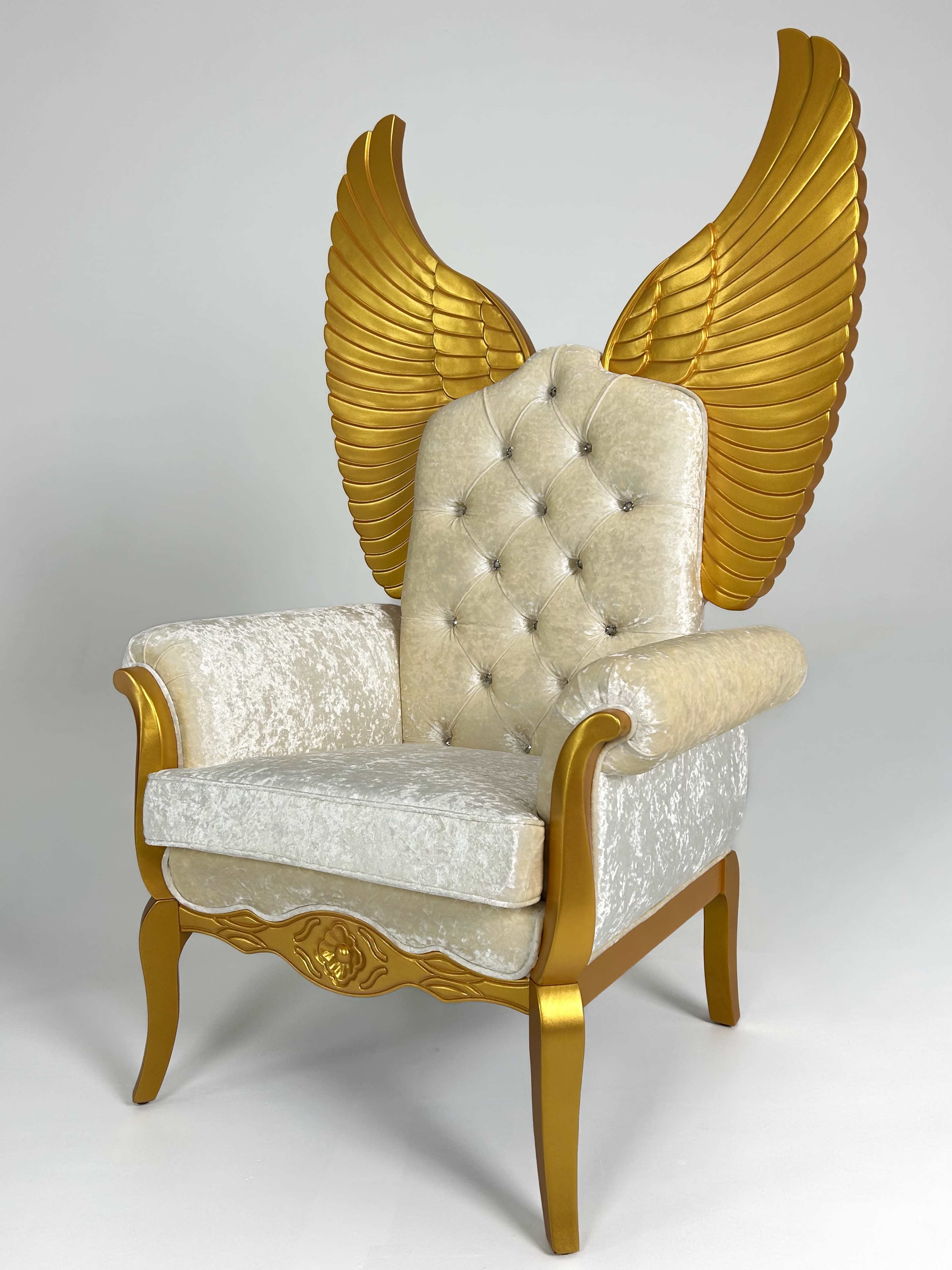 The image shows a decorative armchair with gold wing-like features attached to the backrest, featuring a tufted light-colored upholstery.