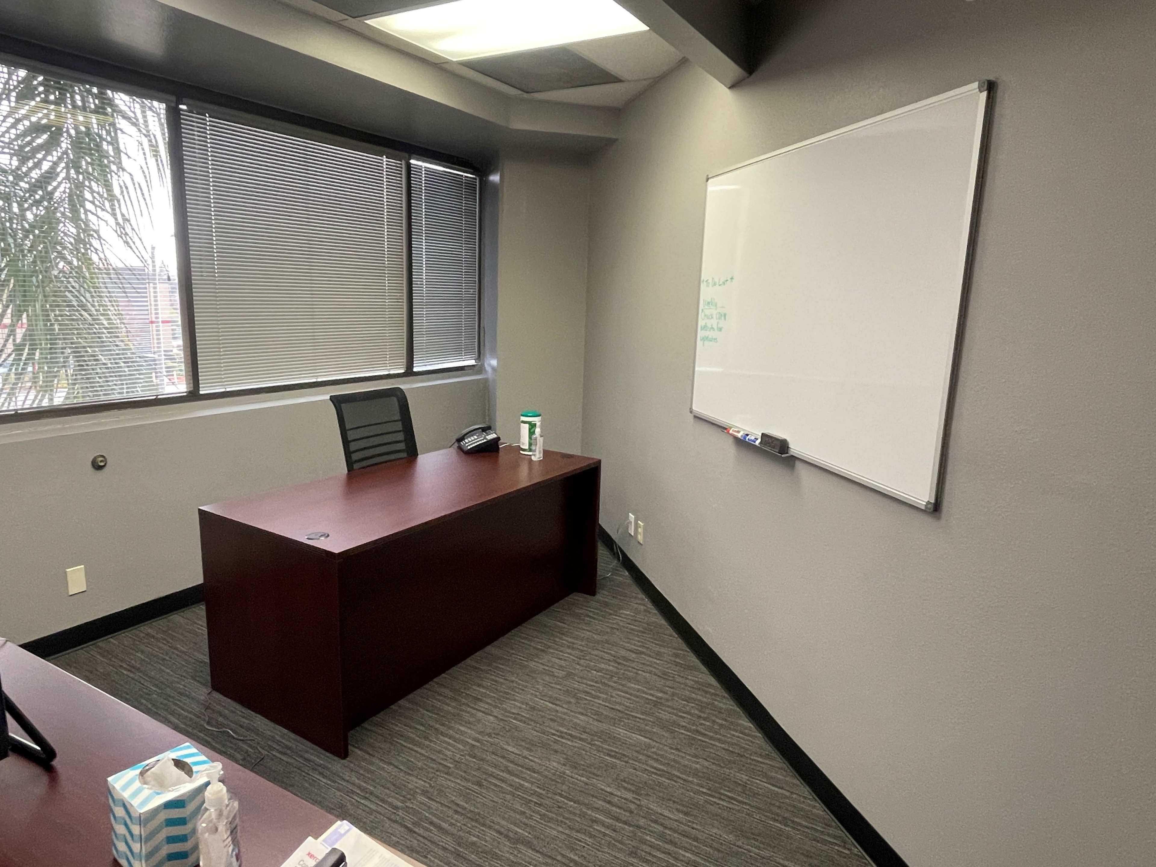 A simple office space featuring a brown desk, a black office chair, a whiteboard on the wall, and a window with horizontal blinds.
