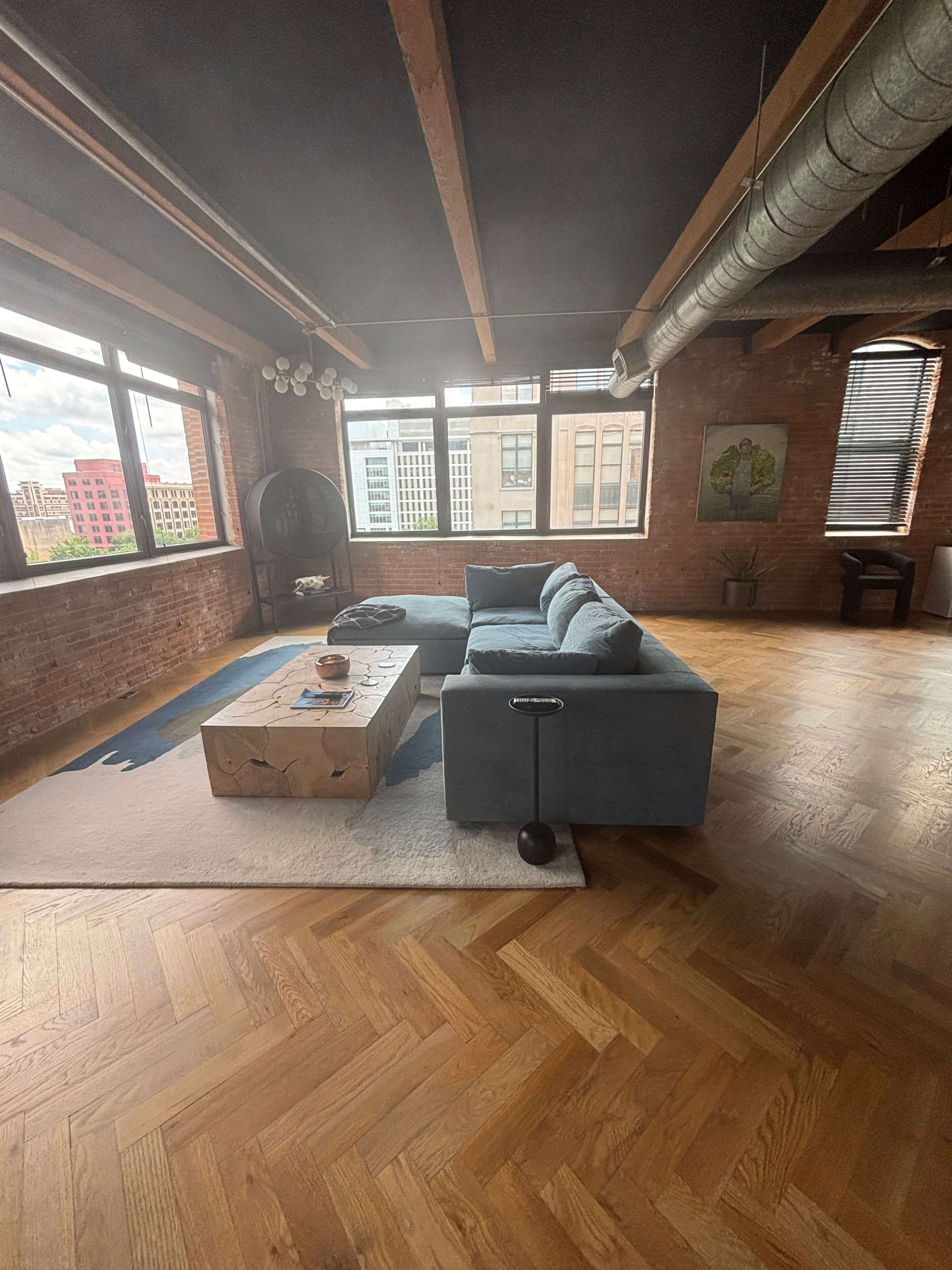 The image shows a modern living room with a blue sectional couch, a wooden coffee table, and large windows that provide a view of the city outside.