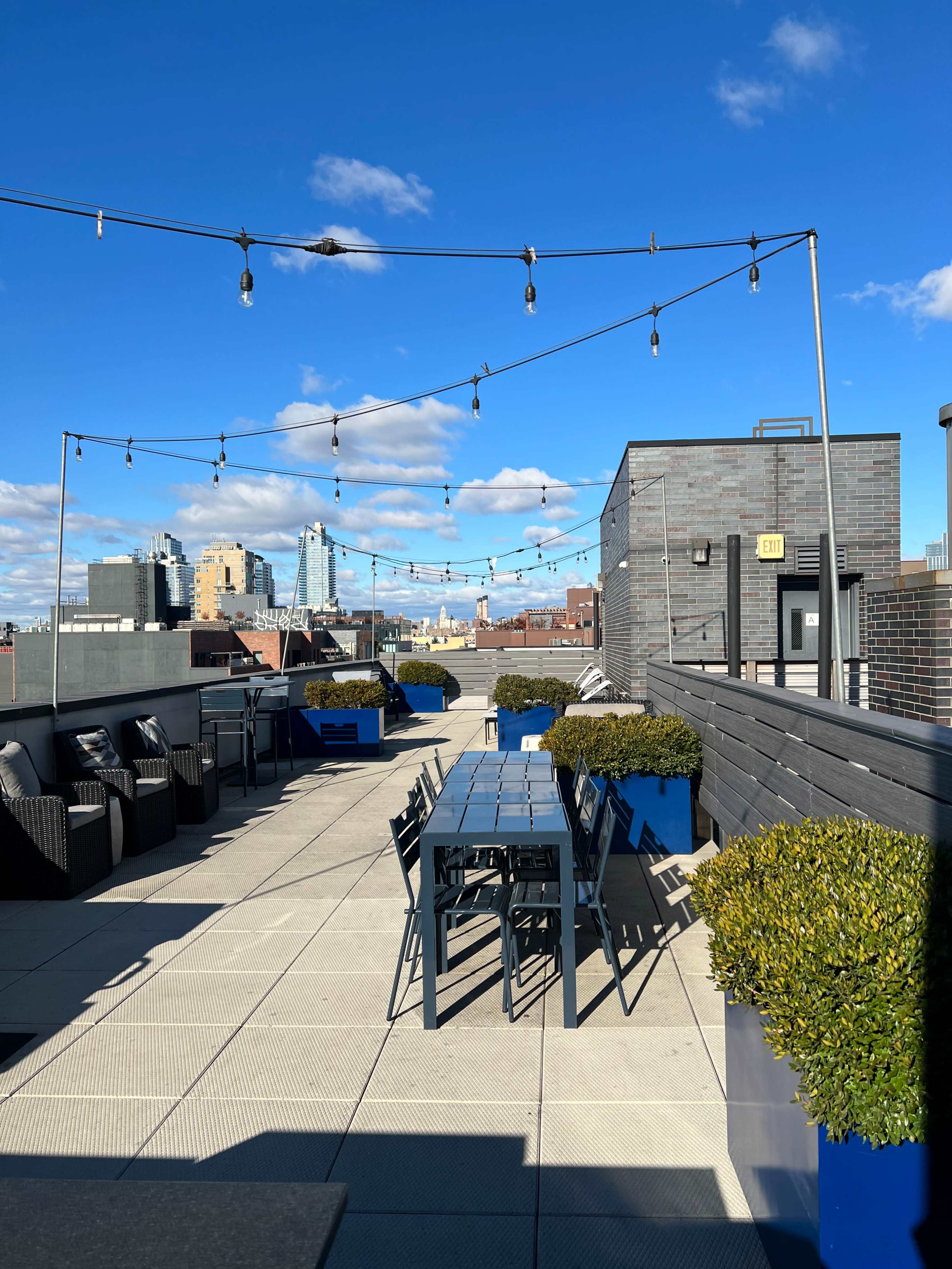 A rooftop terrace with string lights, a long dining table, and planters, set against a blue sky and city skyline.