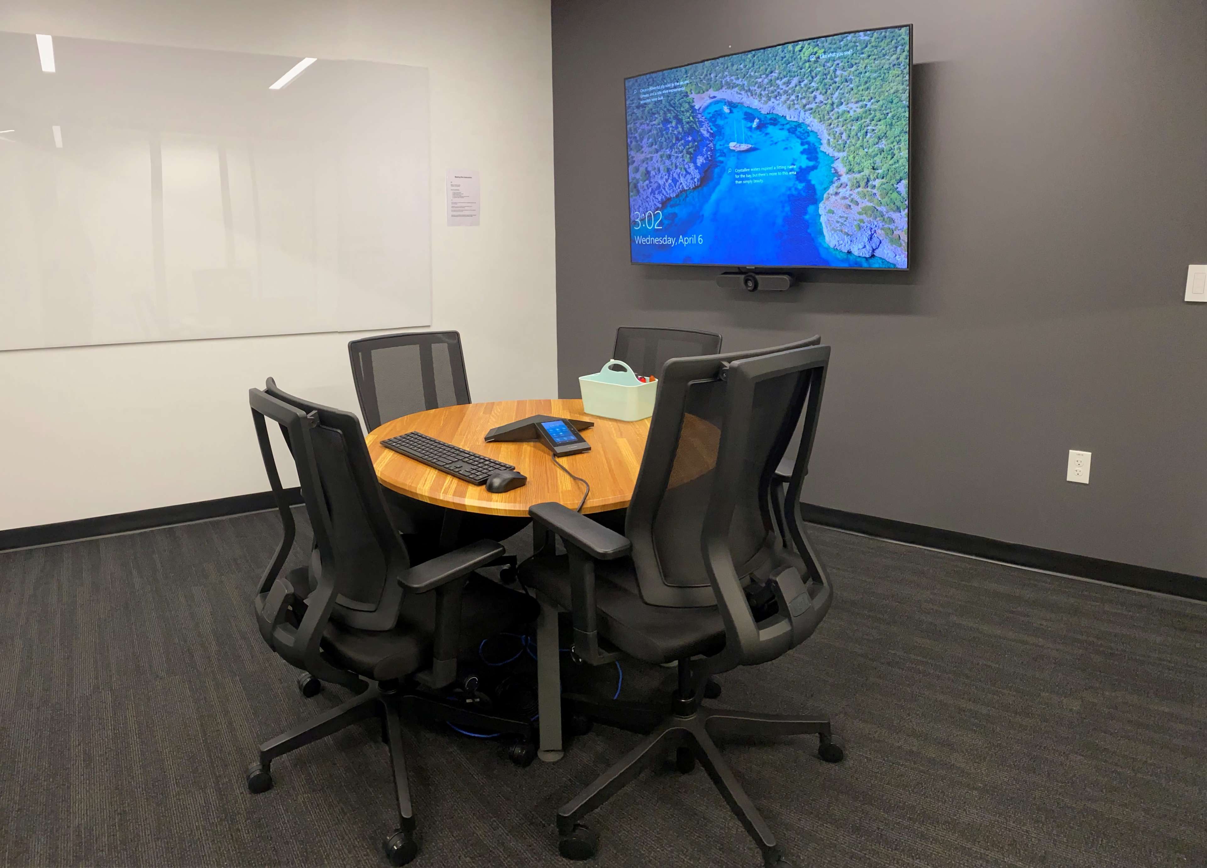 A conference room features a round wooden table surrounded by five ergonomic chairs, with a monitor displaying a scenic landscape mounted on the wall.
