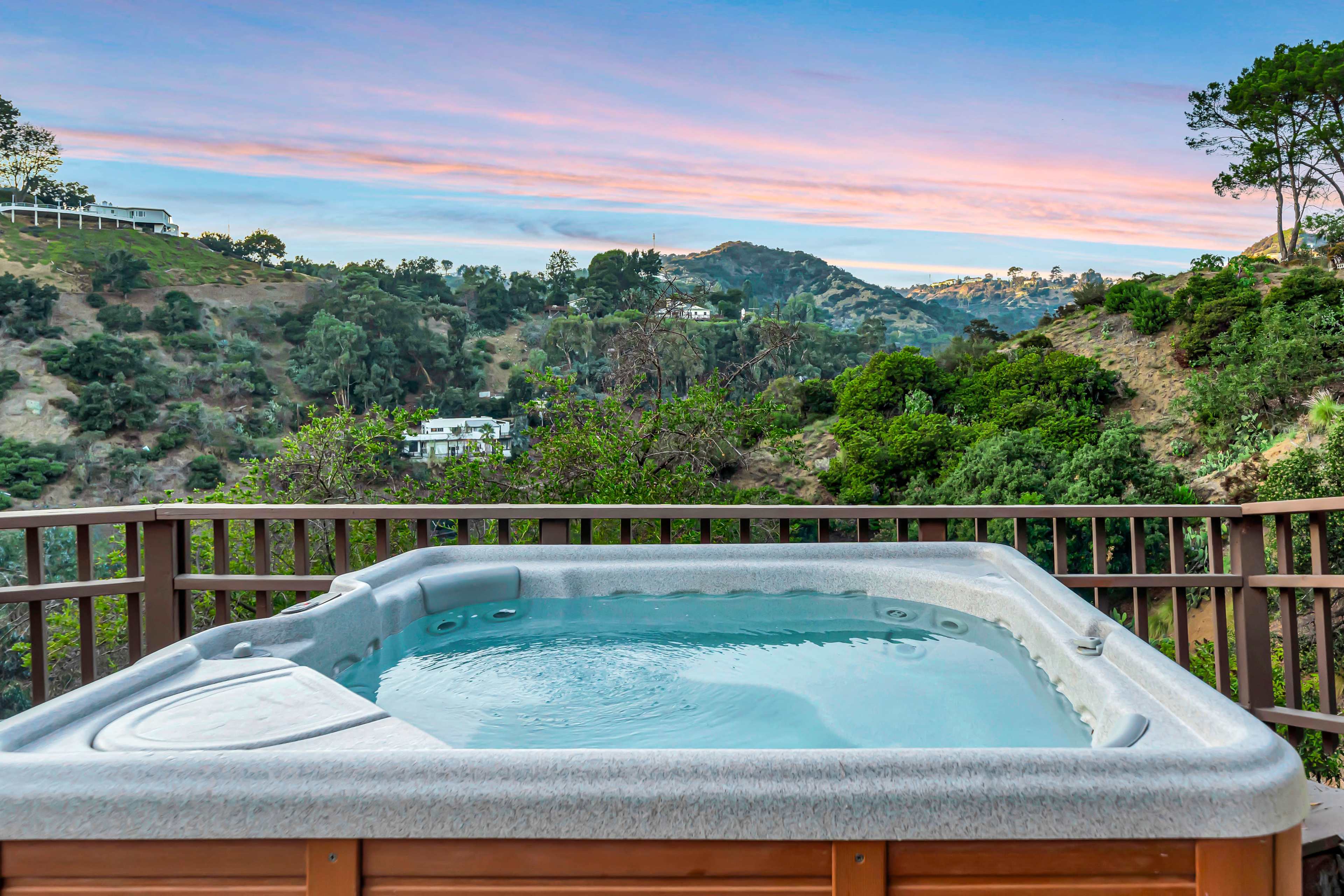 A hot tub overlooks a hillside landscape with trees and distant homes under a colorful sky.