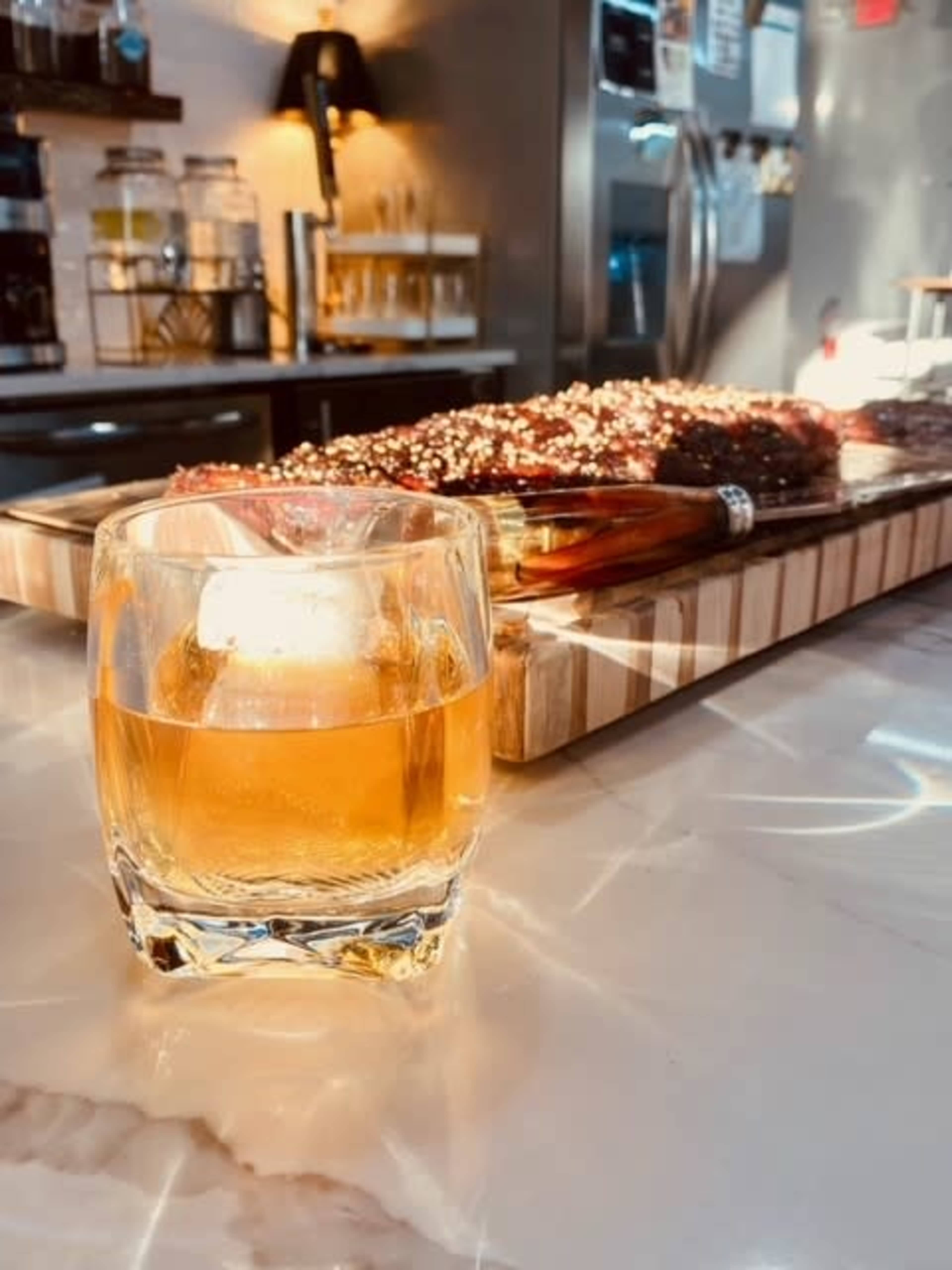 A glass of whiskey sits on a marble counter in the foreground, while a platter of food is visible in the background.