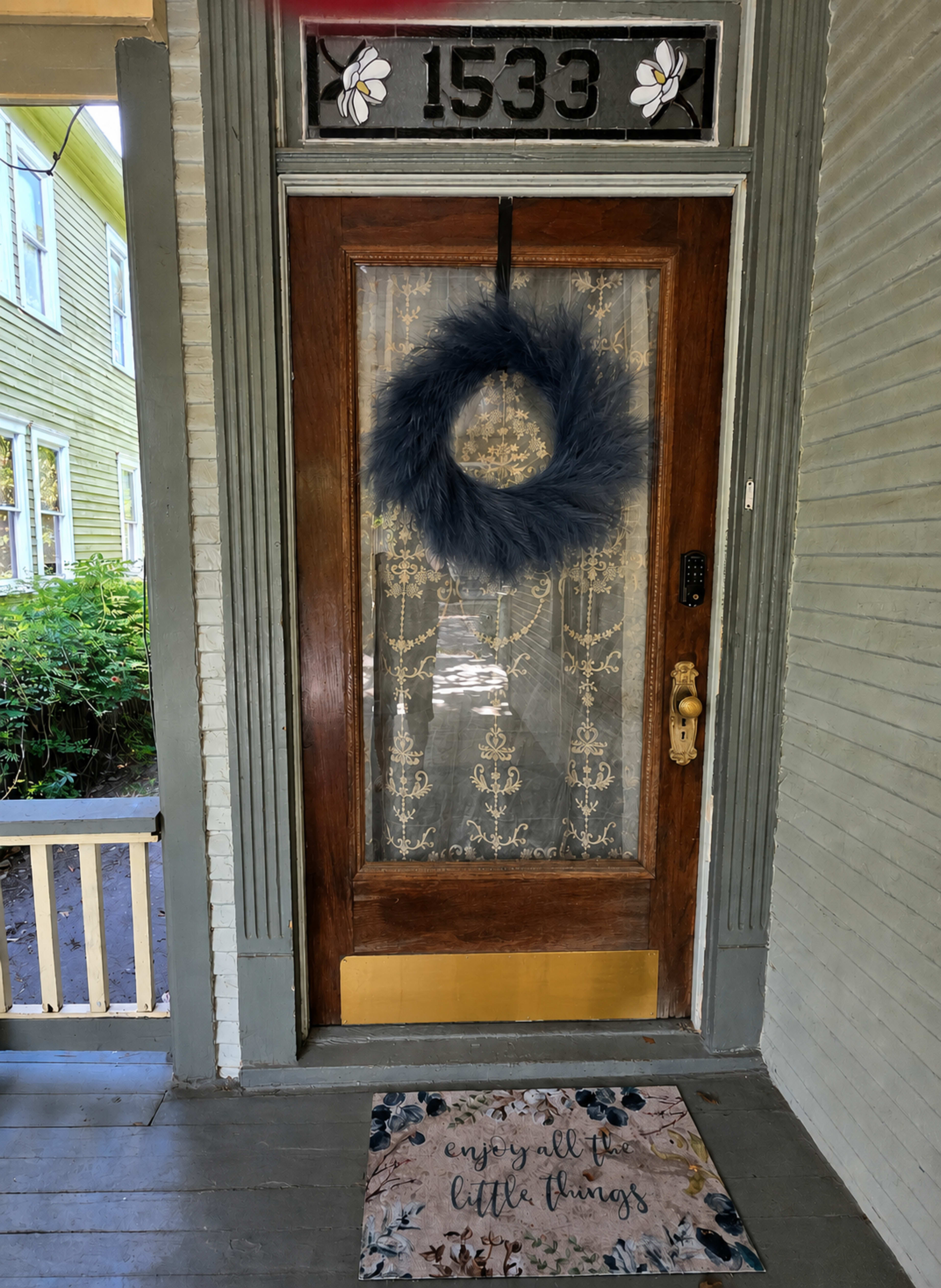 The image shows a wooden front door with a decorative wreath, a welcome mat, and a lace curtain behind glass.
