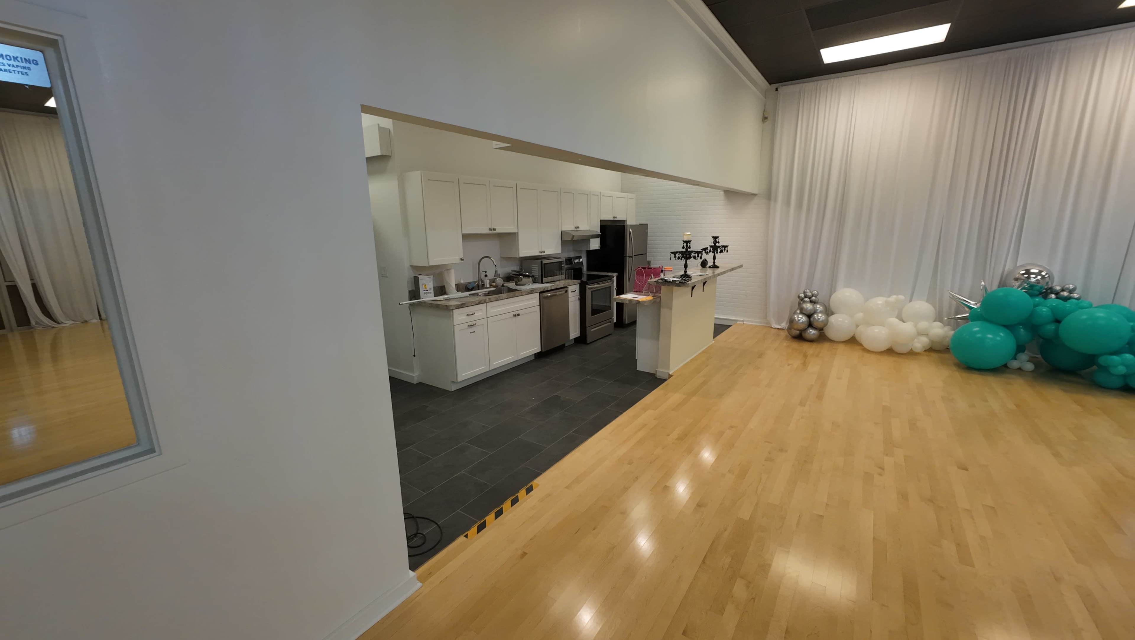 A spacious room with a kitchen area featuring white cabinets and stainless steel appliances, and a corner decorated with balloons in teal and white.