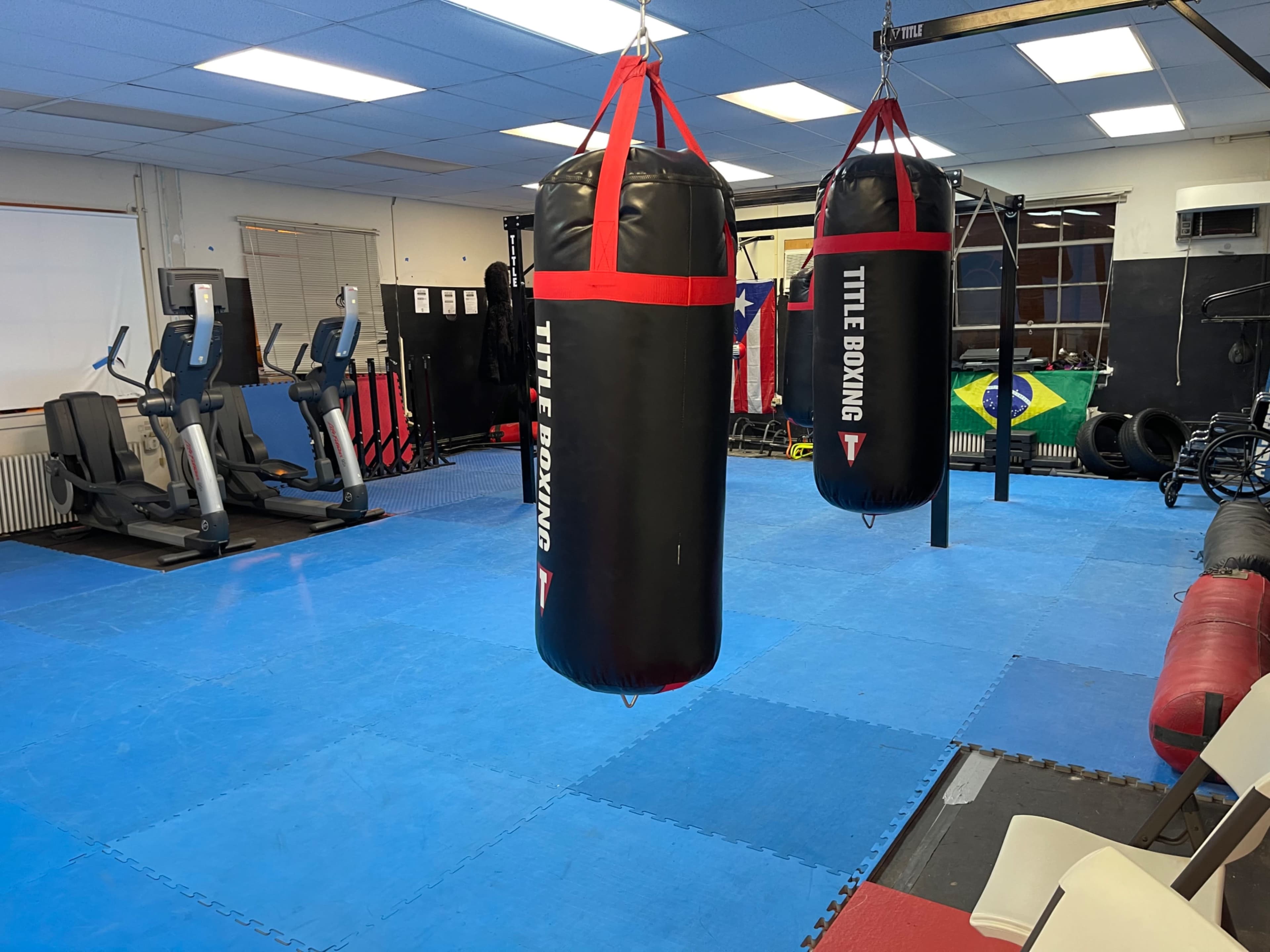 A boxing gym with two heavy bags hanging from the ceiling, exercise equipment on the left, and a blue mat flooring.
