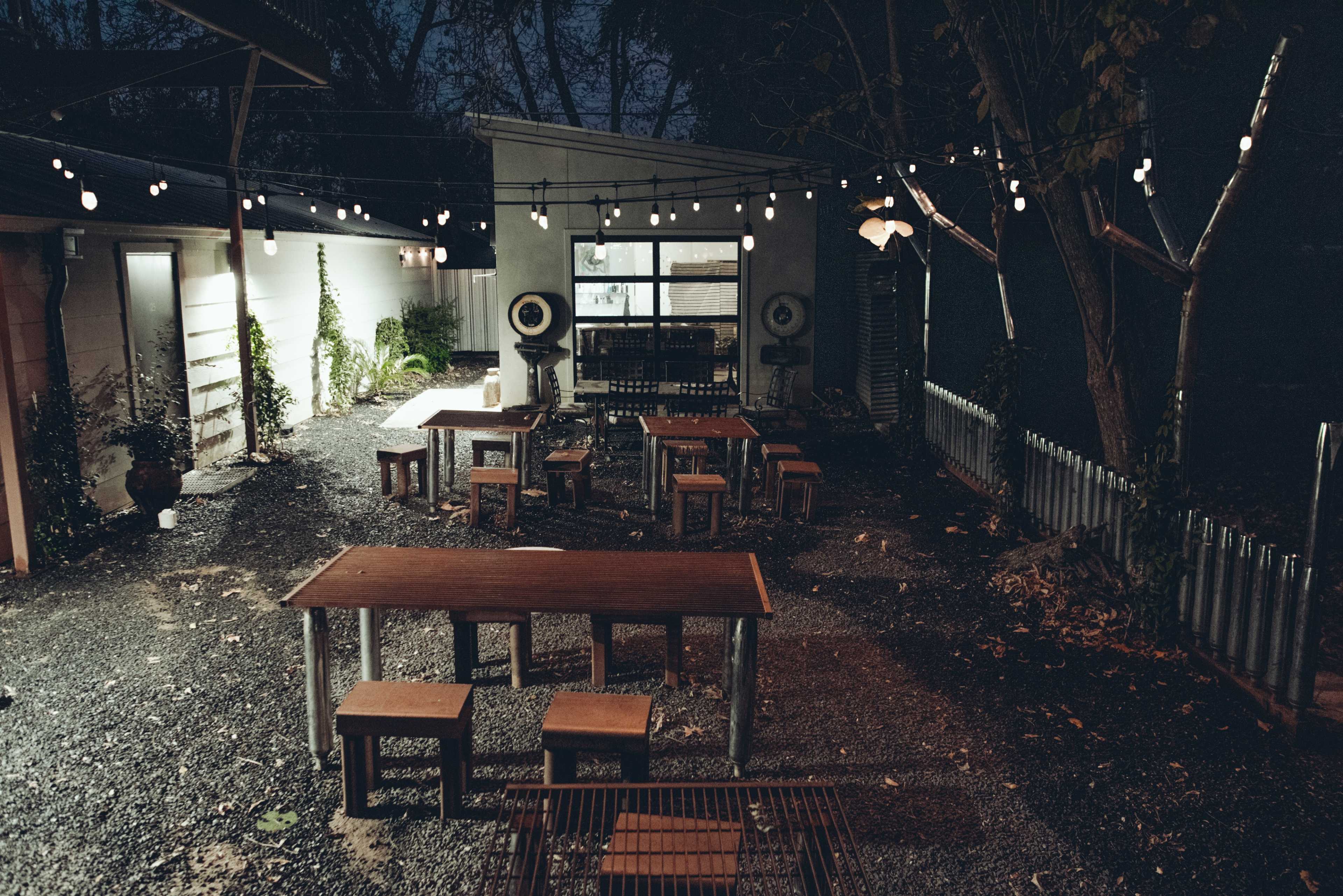 The image shows an outdoor dining area at night, featuring several wooden tables and benches surrounded by string lights and greenery.