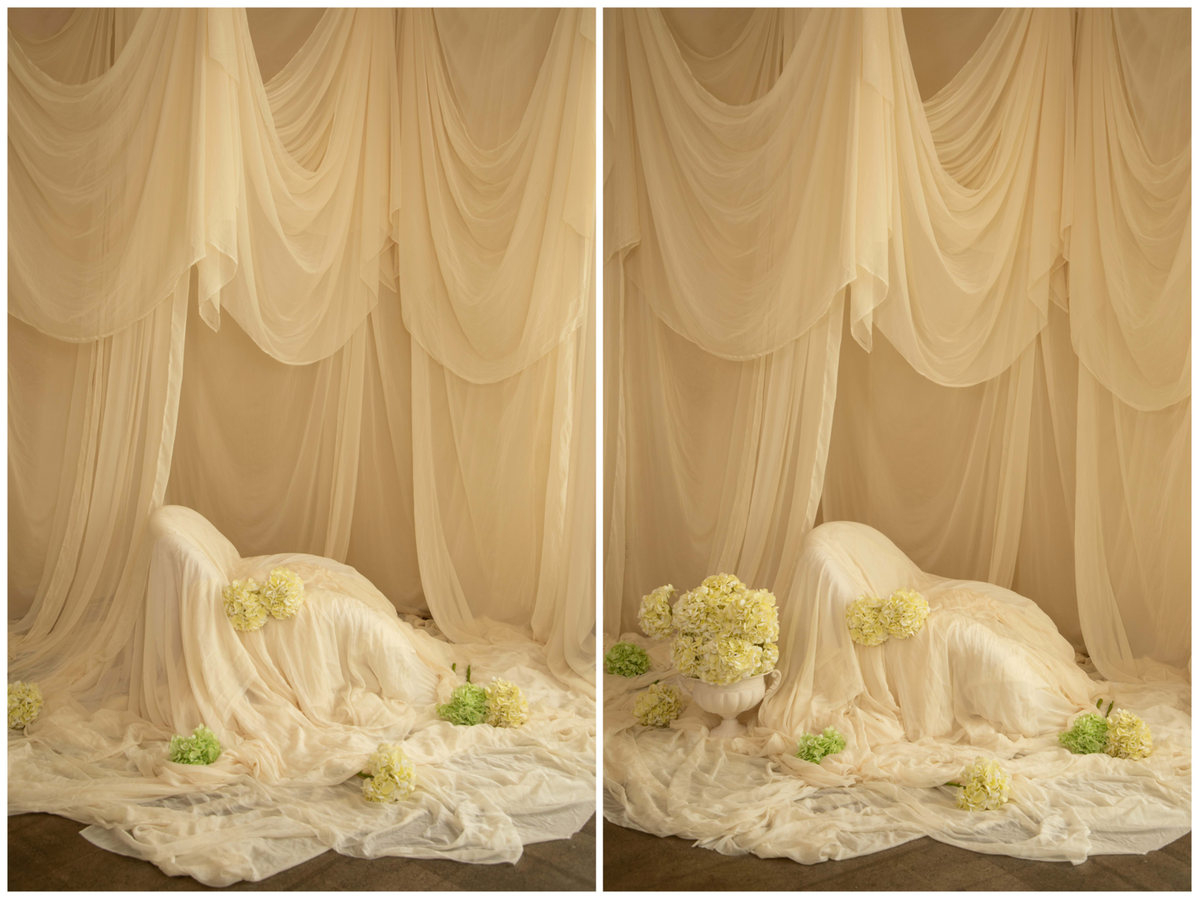 The image features a softly draped setup with flowing beige fabric, showcasing a simple arrangement of pale yellow flowers and green accents on the floor.