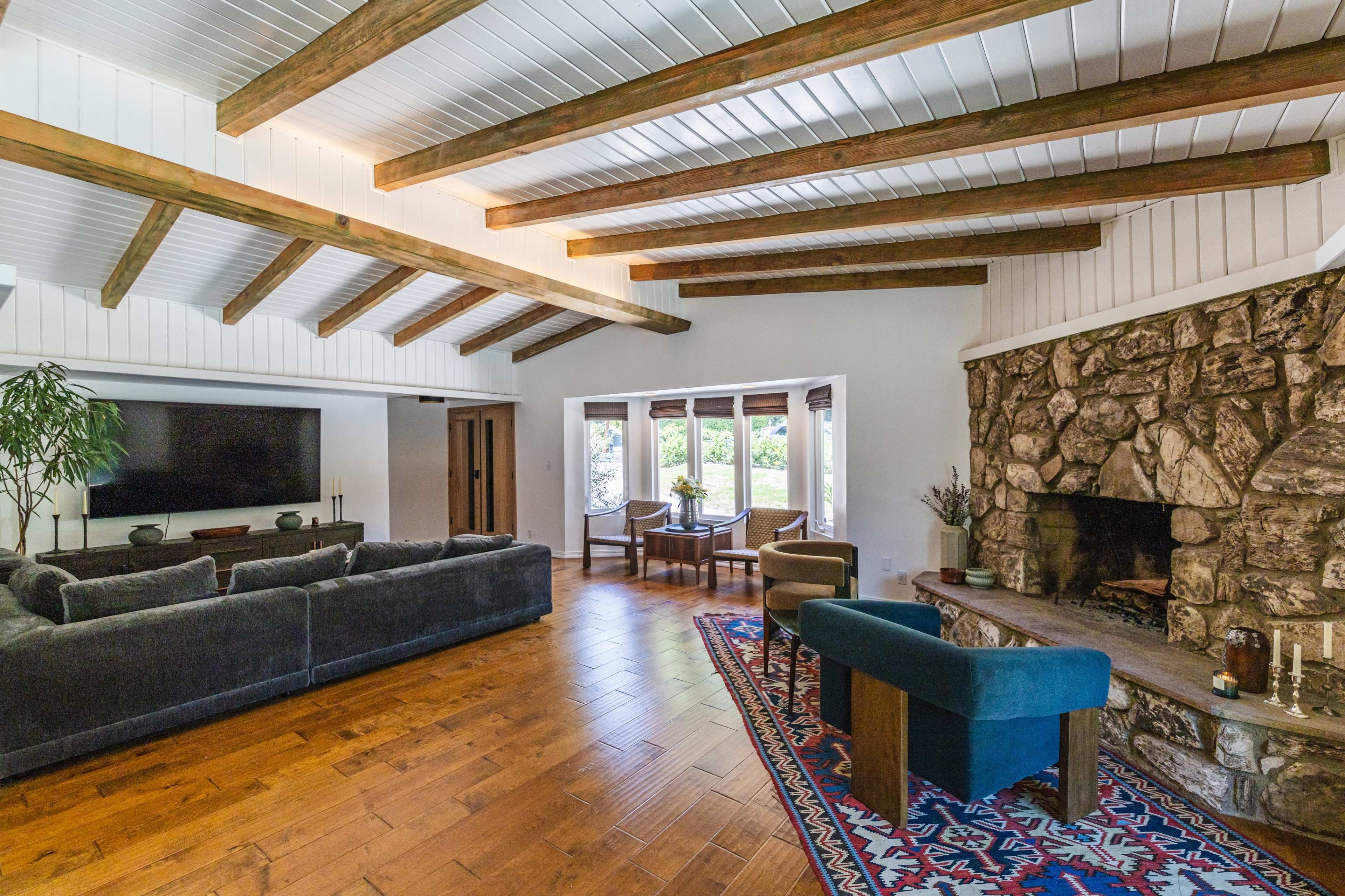 A spacious living room features exposed wooden beams, a stone fireplace, a large television, and a seating area with a rug and a coffee table.