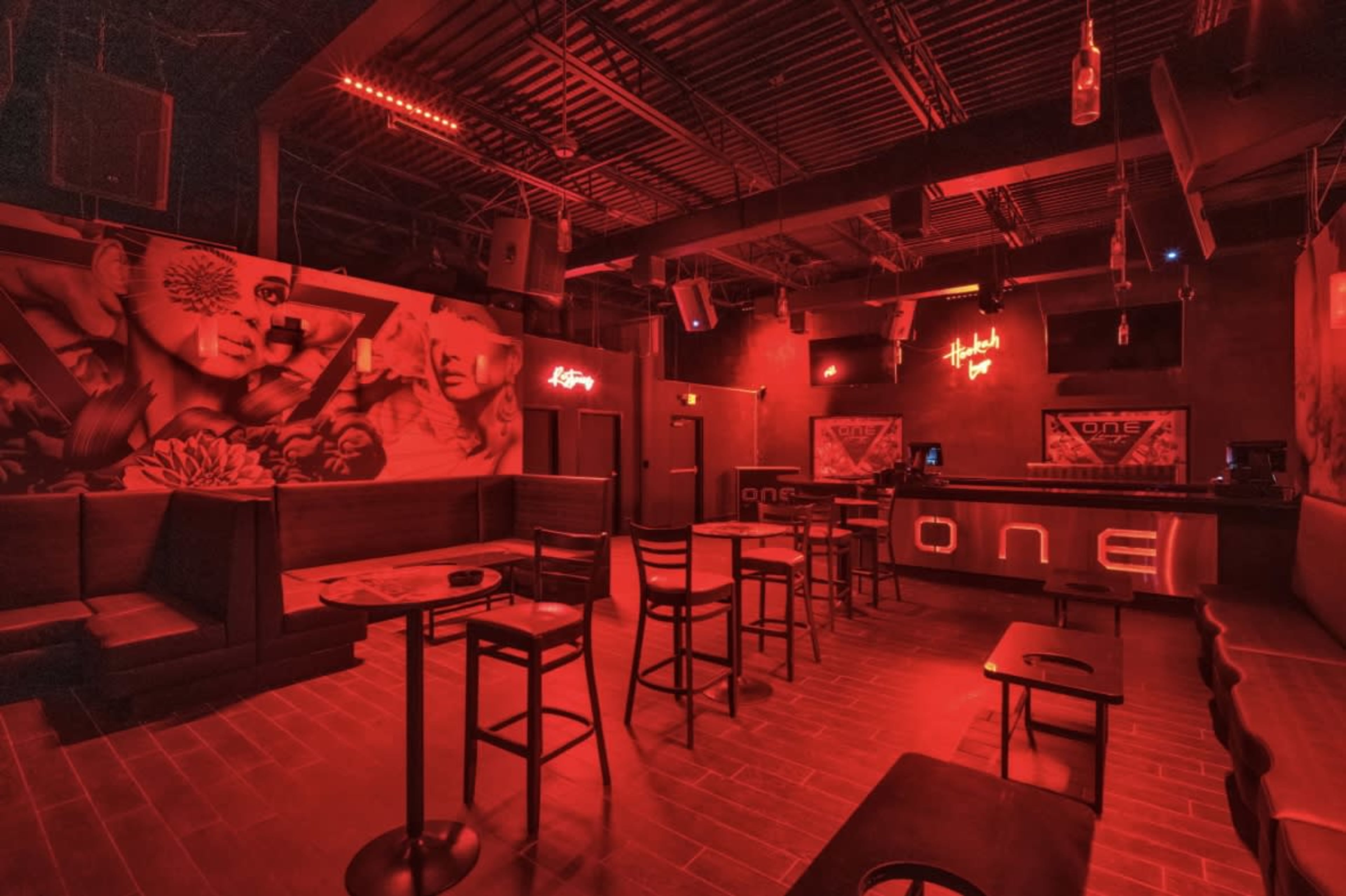 The image shows a dimly lit bar with red lighting, featuring empty seating areas and a bar counter with neon signage.