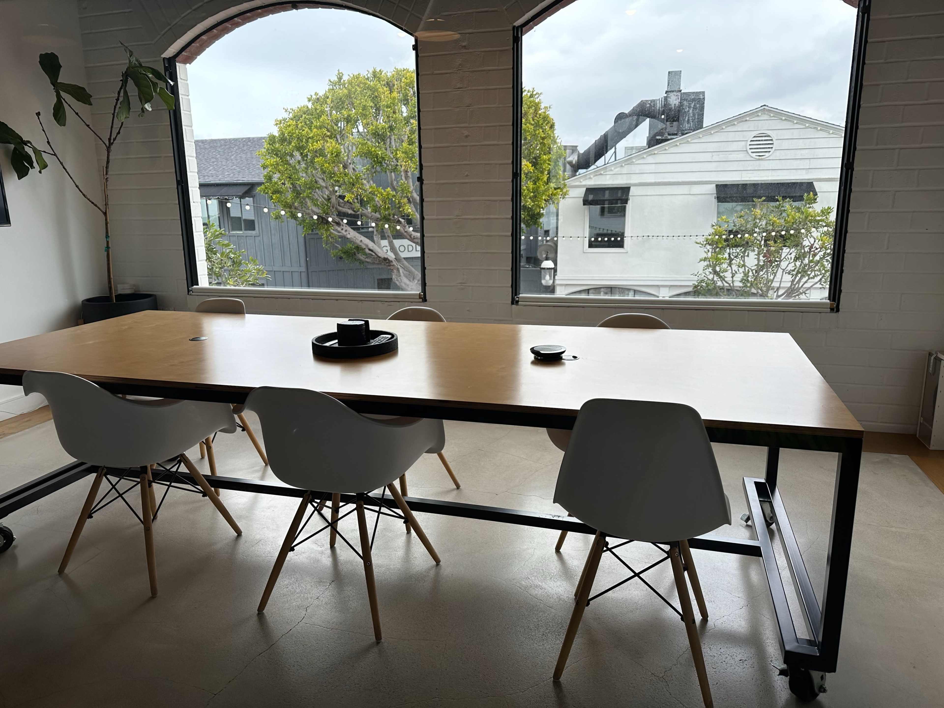 A modern meeting room features a long wooden table surrounded by white chairs, with large windows displaying a view of buildings and greenery outside.