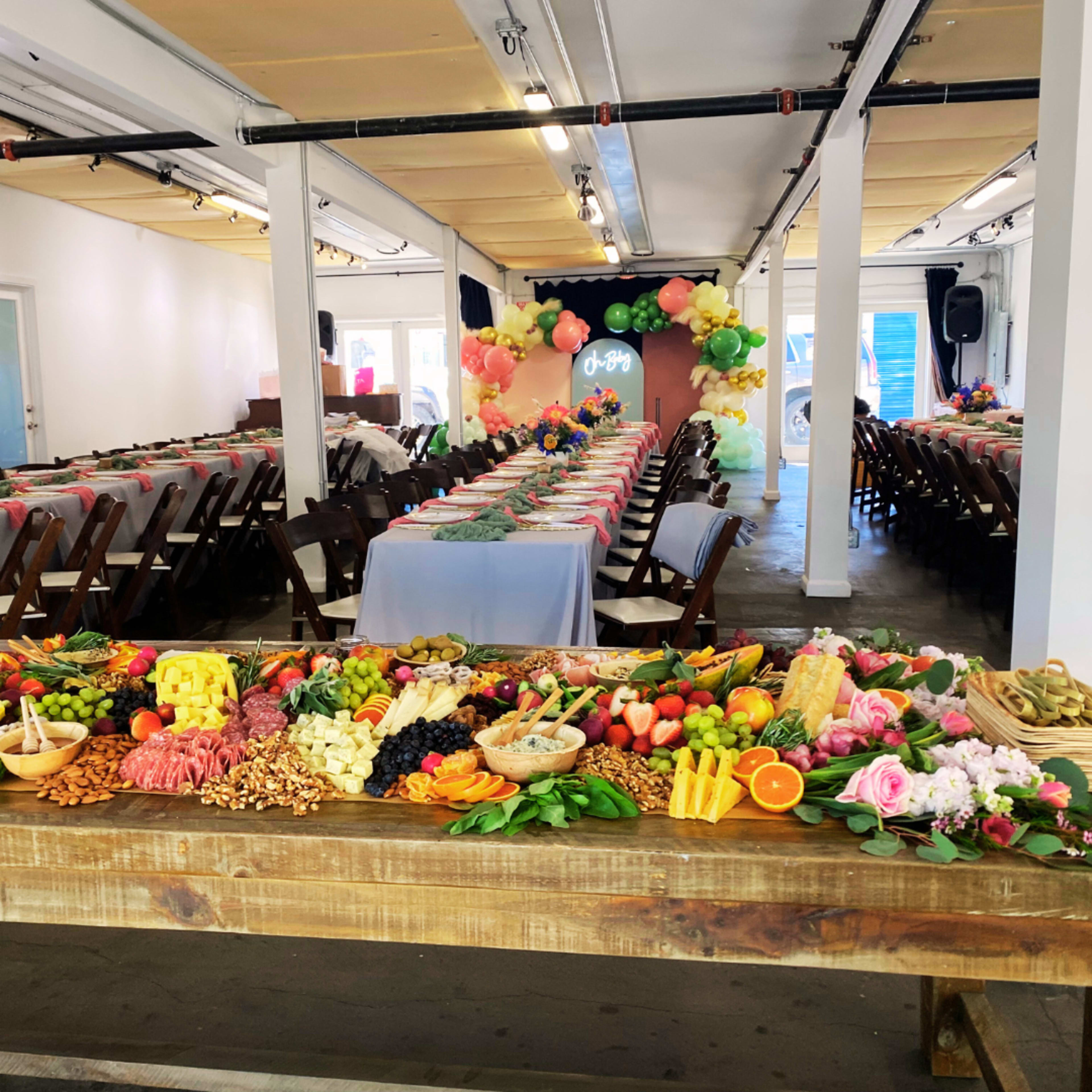A long table displays an elaborate spread of fruits, cheeses, meats, and decorative flowers in a brightly lit event space.