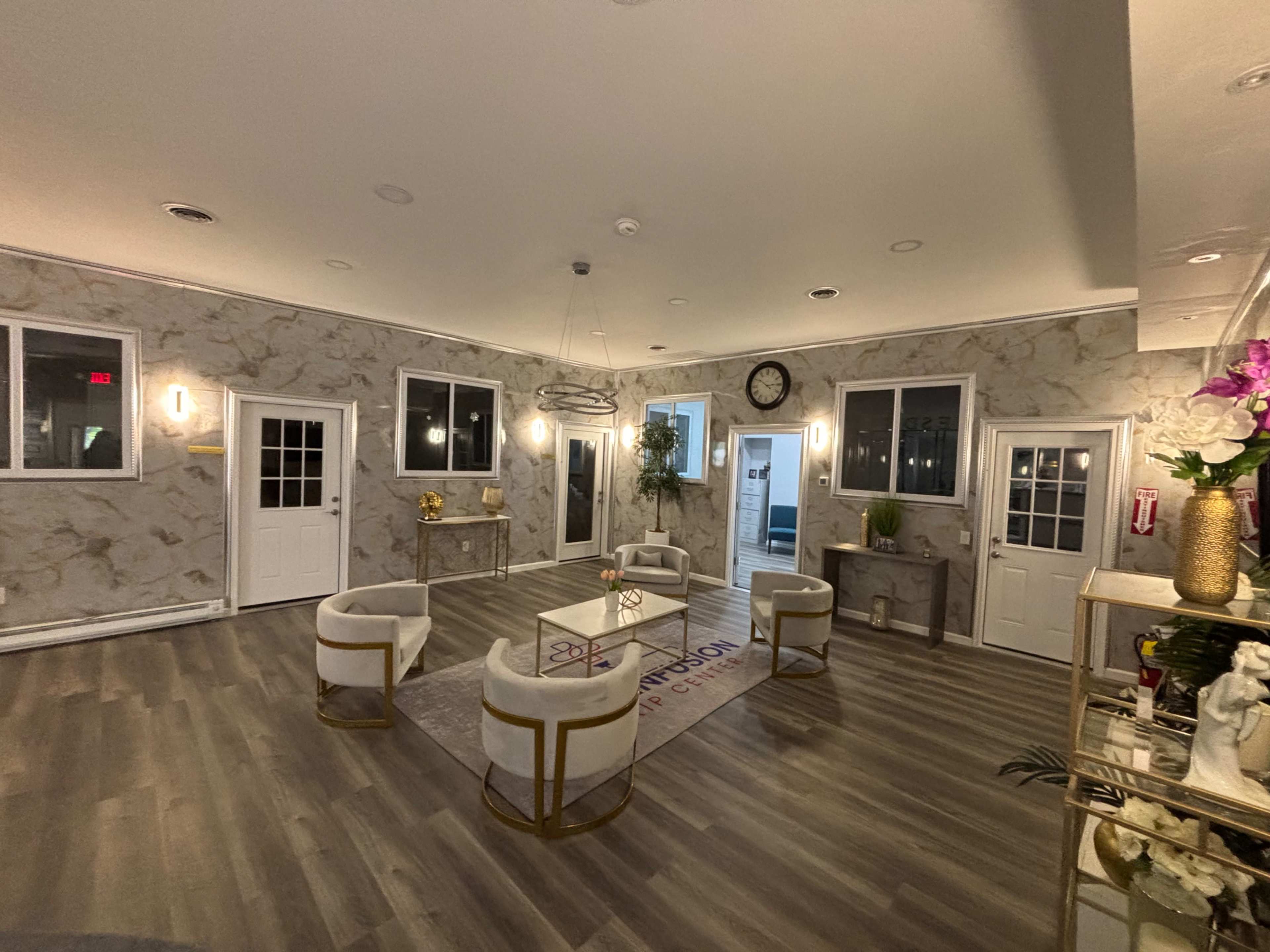 The image shows a well-lit reception area with marble-patterned walls, gray wooden flooring, and several seating arrangements consisting of white chairs around a circular table.