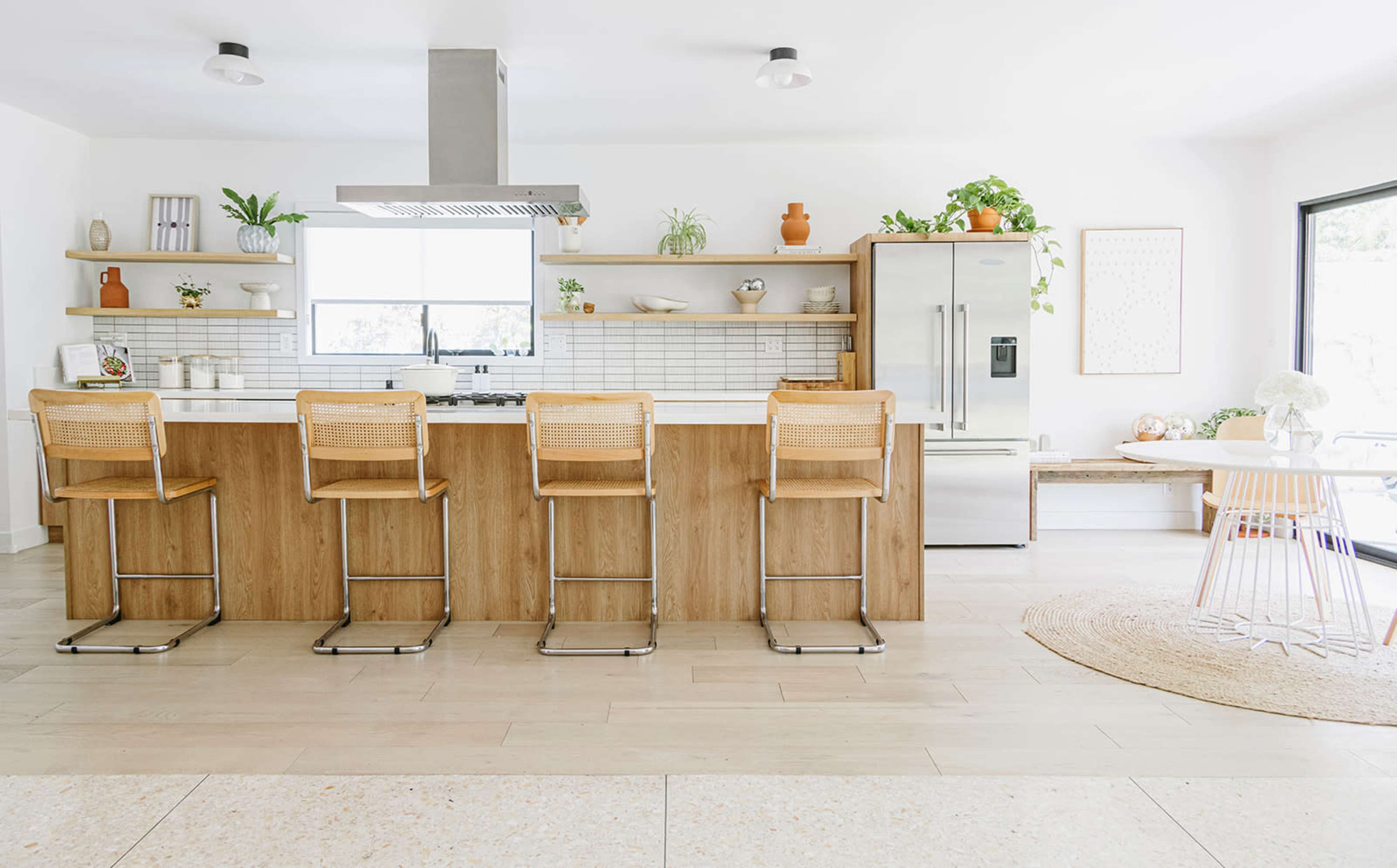 A modern kitchen features a central island with four wooden bar stools, stainless steel appliances, and open shelves decorated with plants and dishware.