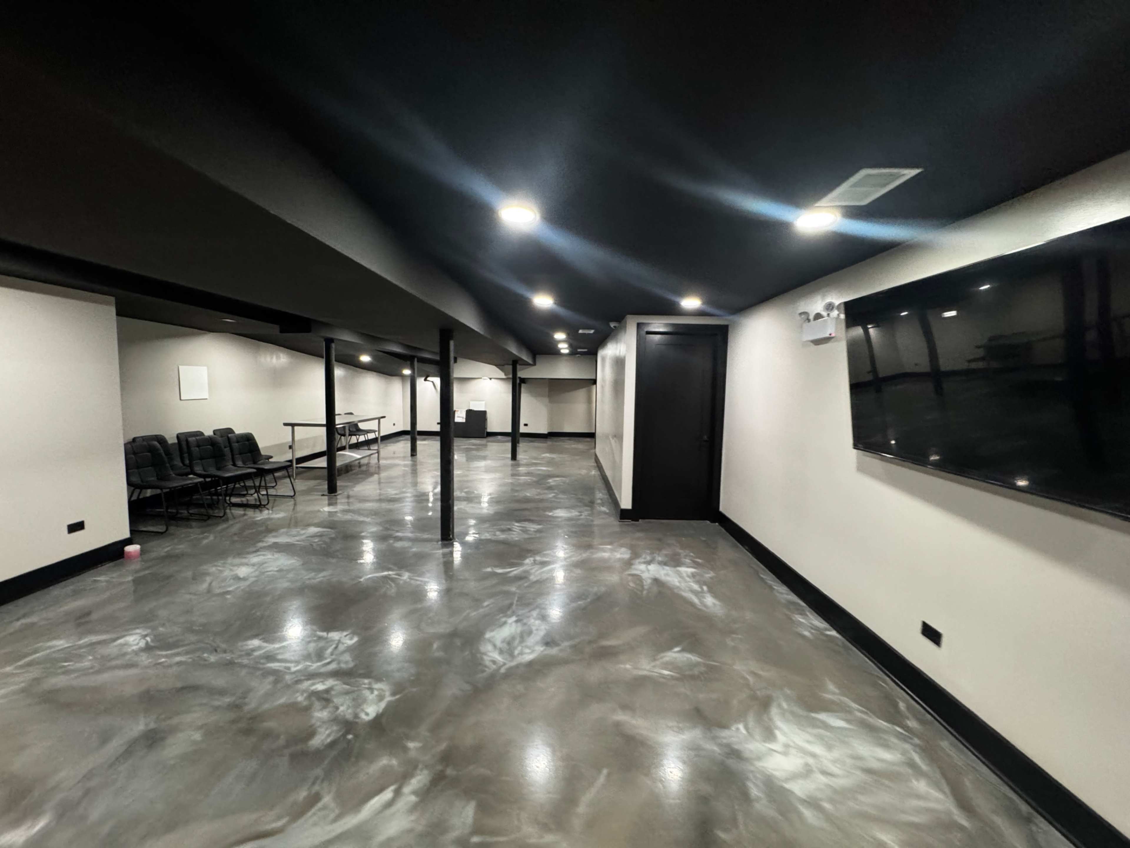 The image shows a spacious, modern basement area with polished concrete flooring, low lighting, and a wall-mounted television.