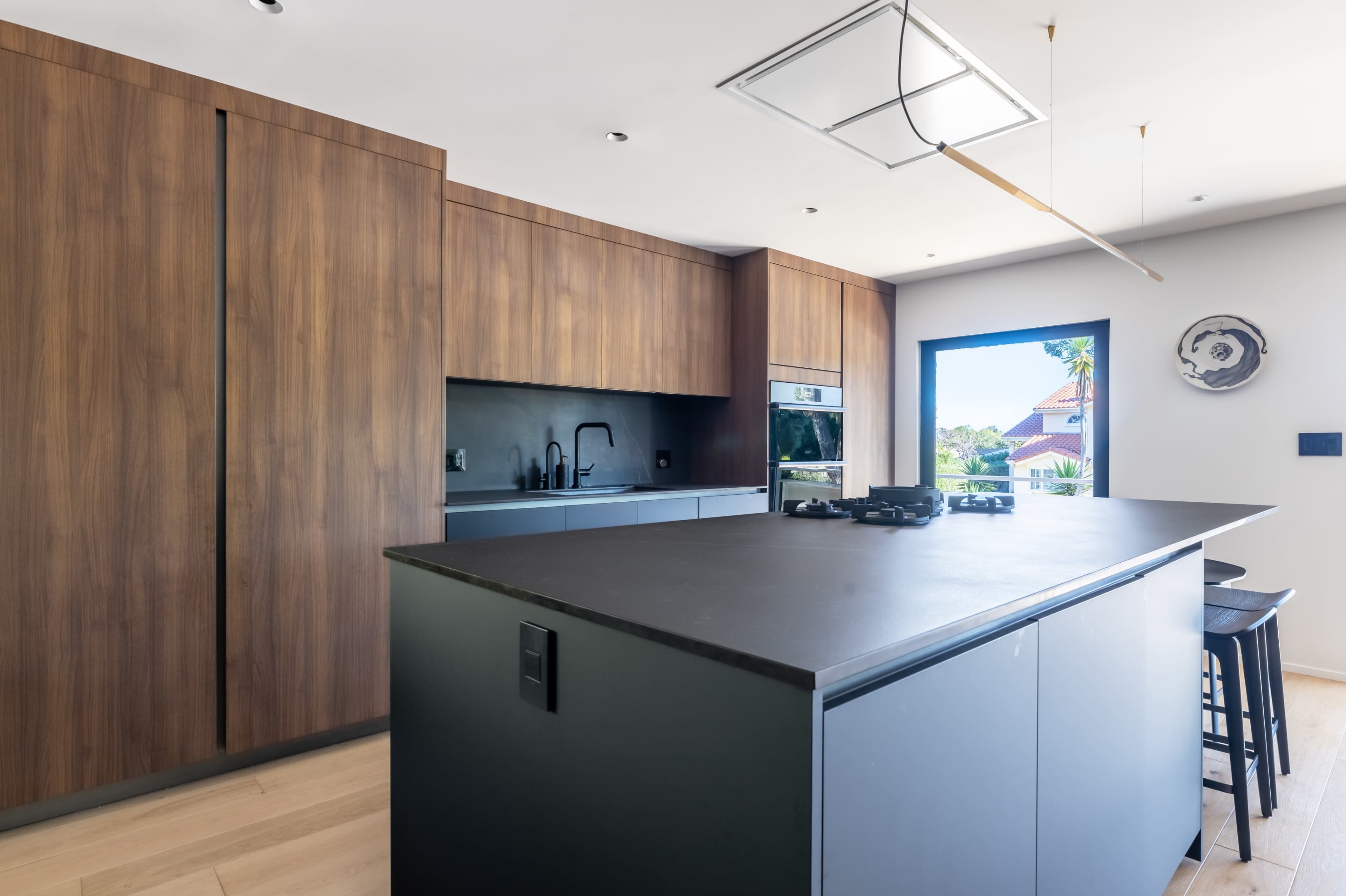 A modern kitchen featuring wooden cabinetry, a central island with a dark countertop, and large windows allowing natural light.