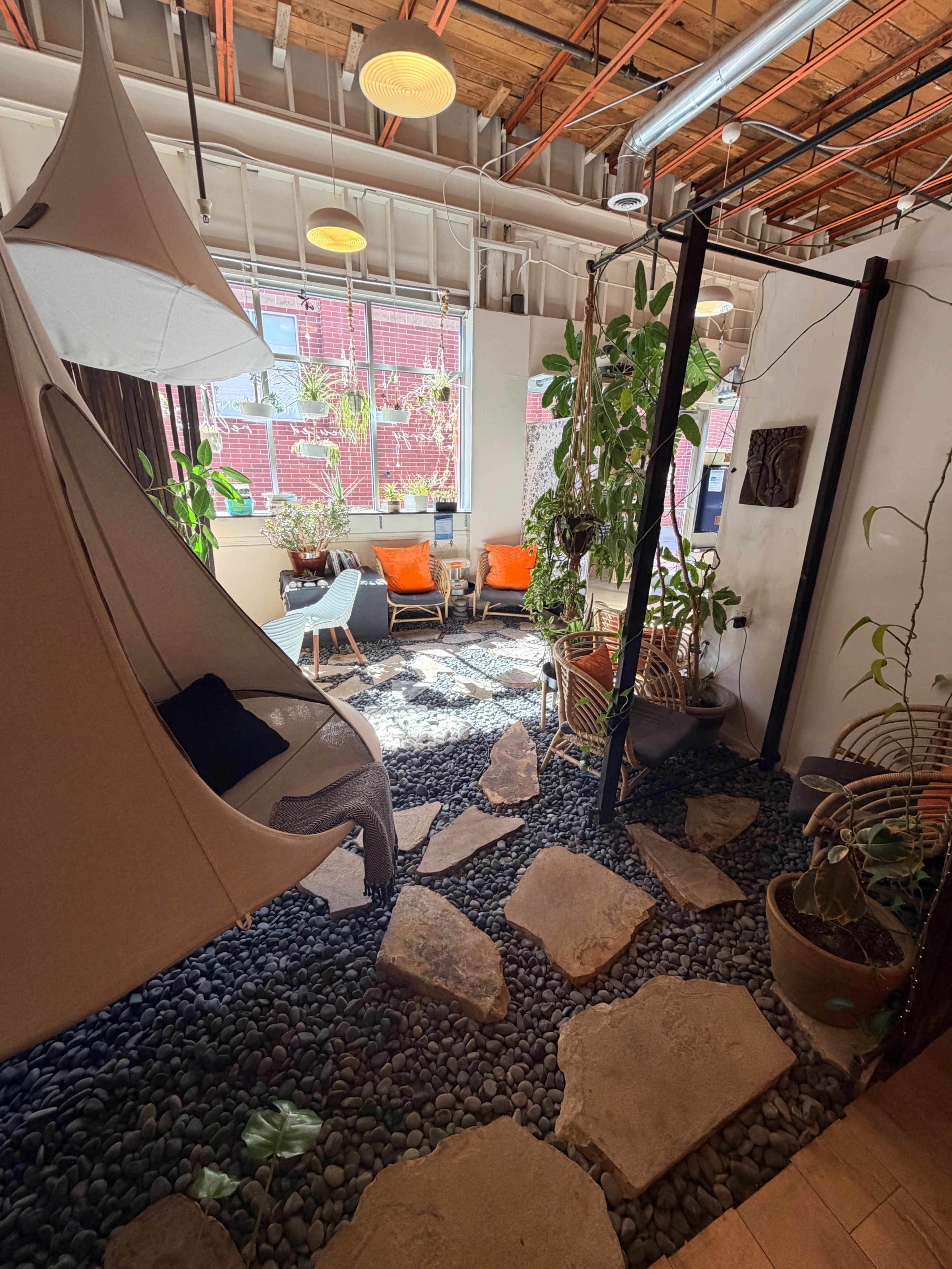 The image shows a modern indoor space with a stone and pebble floor, two orange chairs, a hanging chair, and several indoor plants.