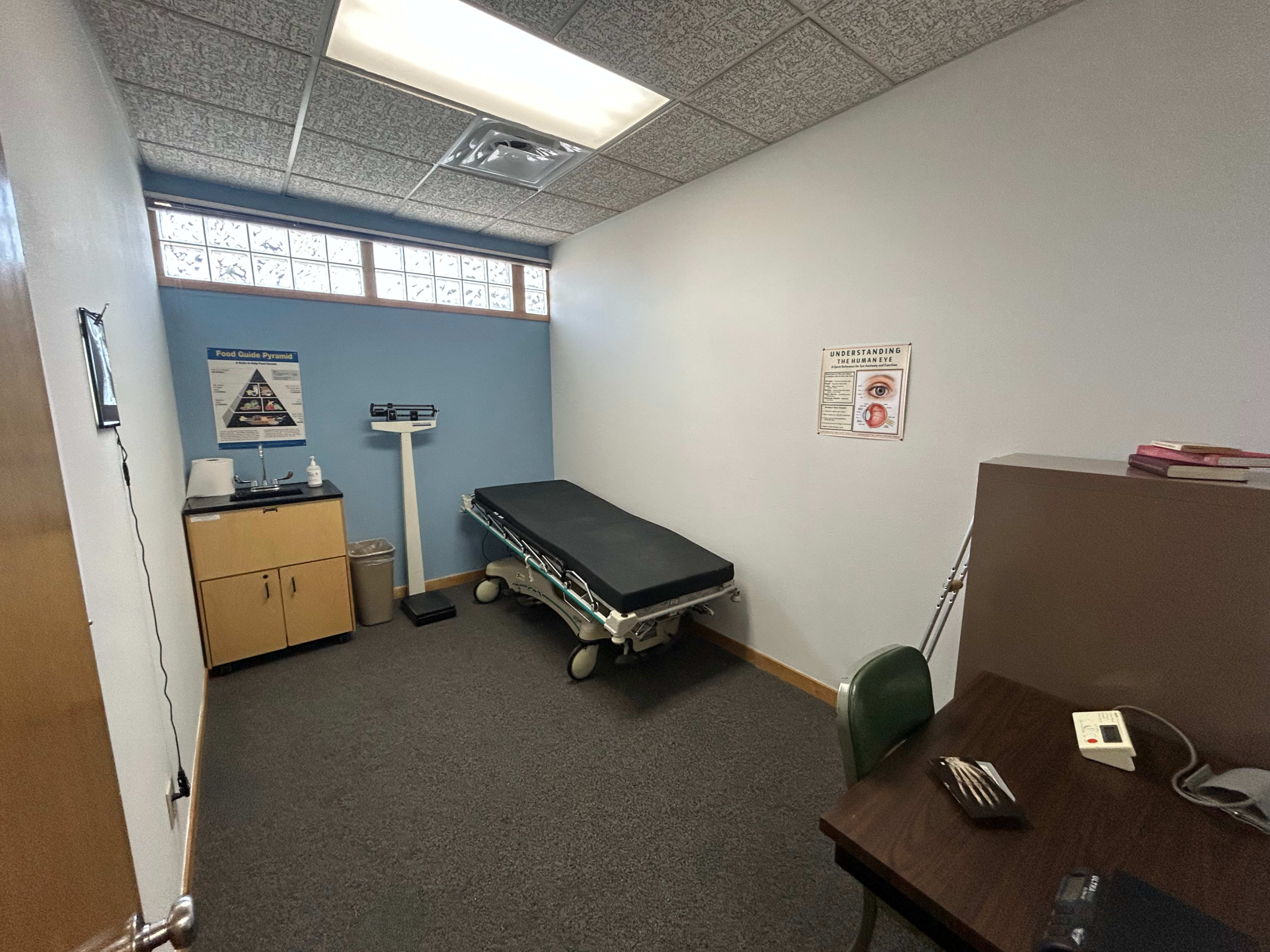 The image shows a small examination room featuring a medical examination table, a scale, a desk with a chair, and a waste bin.