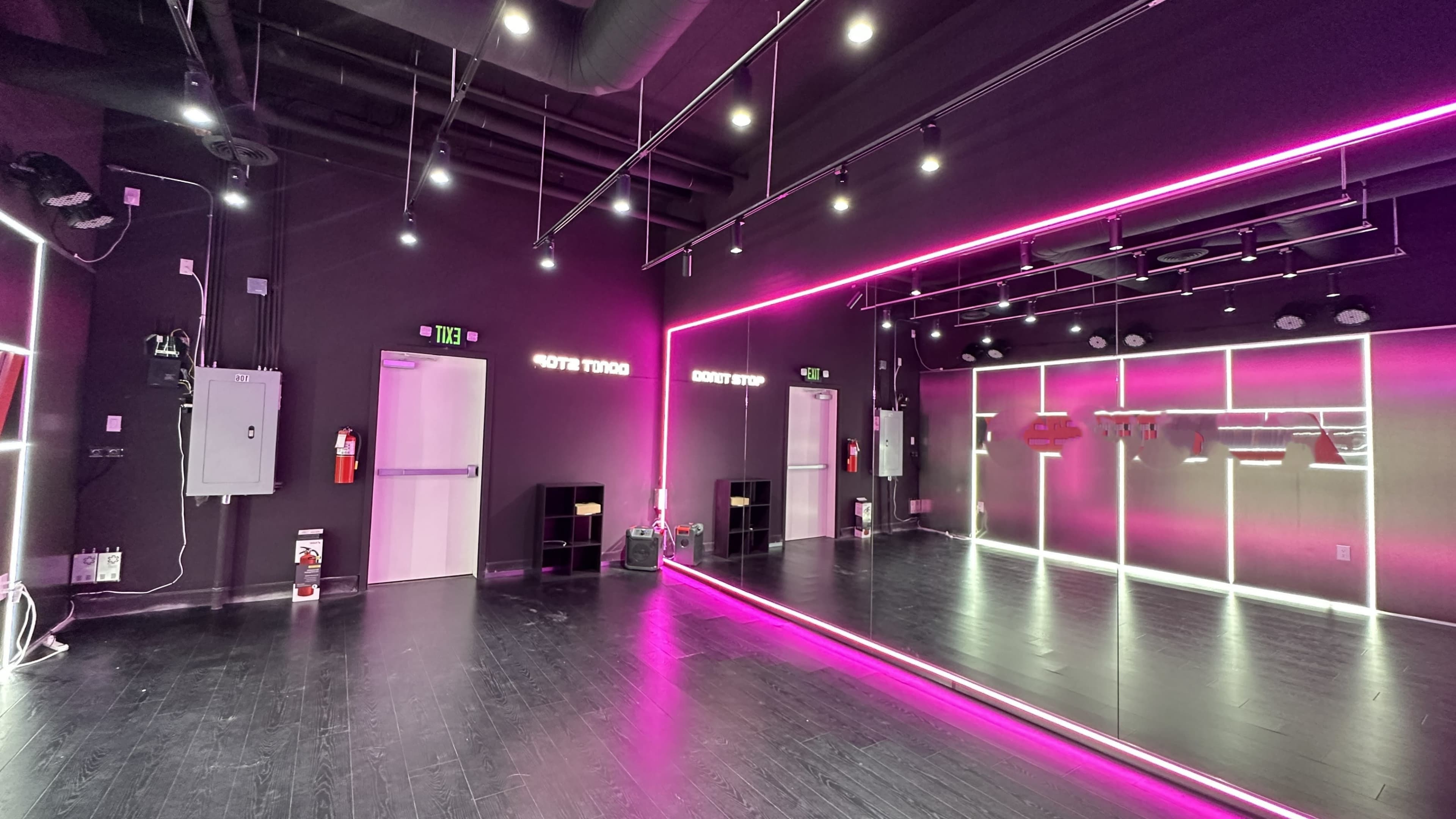 A dance studio featuring black walls, hardwood flooring, and pink neon lights outlining the mirrors and spaces.