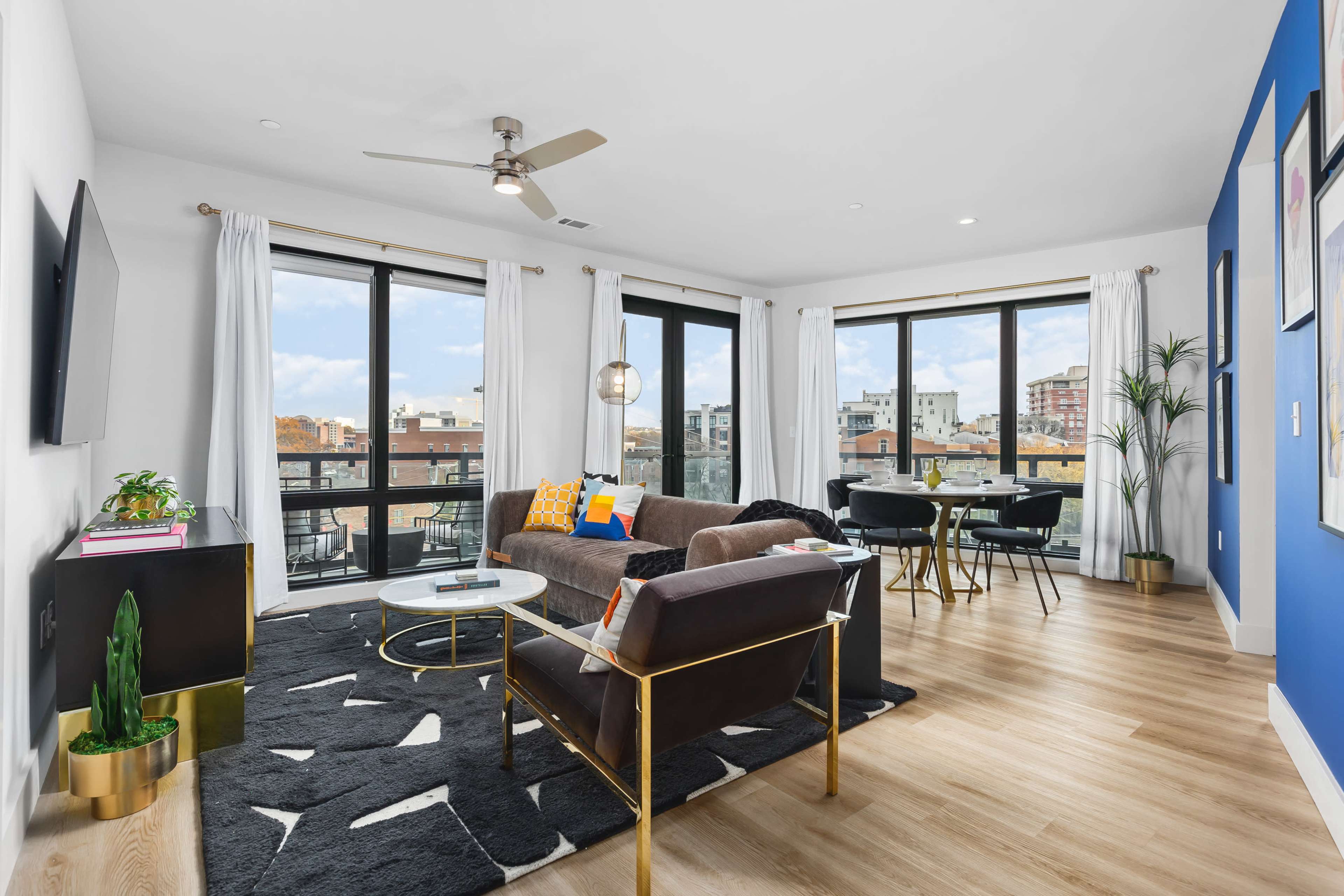A modern living room features a brown sofa, a glass coffee table, and large windows with city views, accented by a dining area and indoor plants.