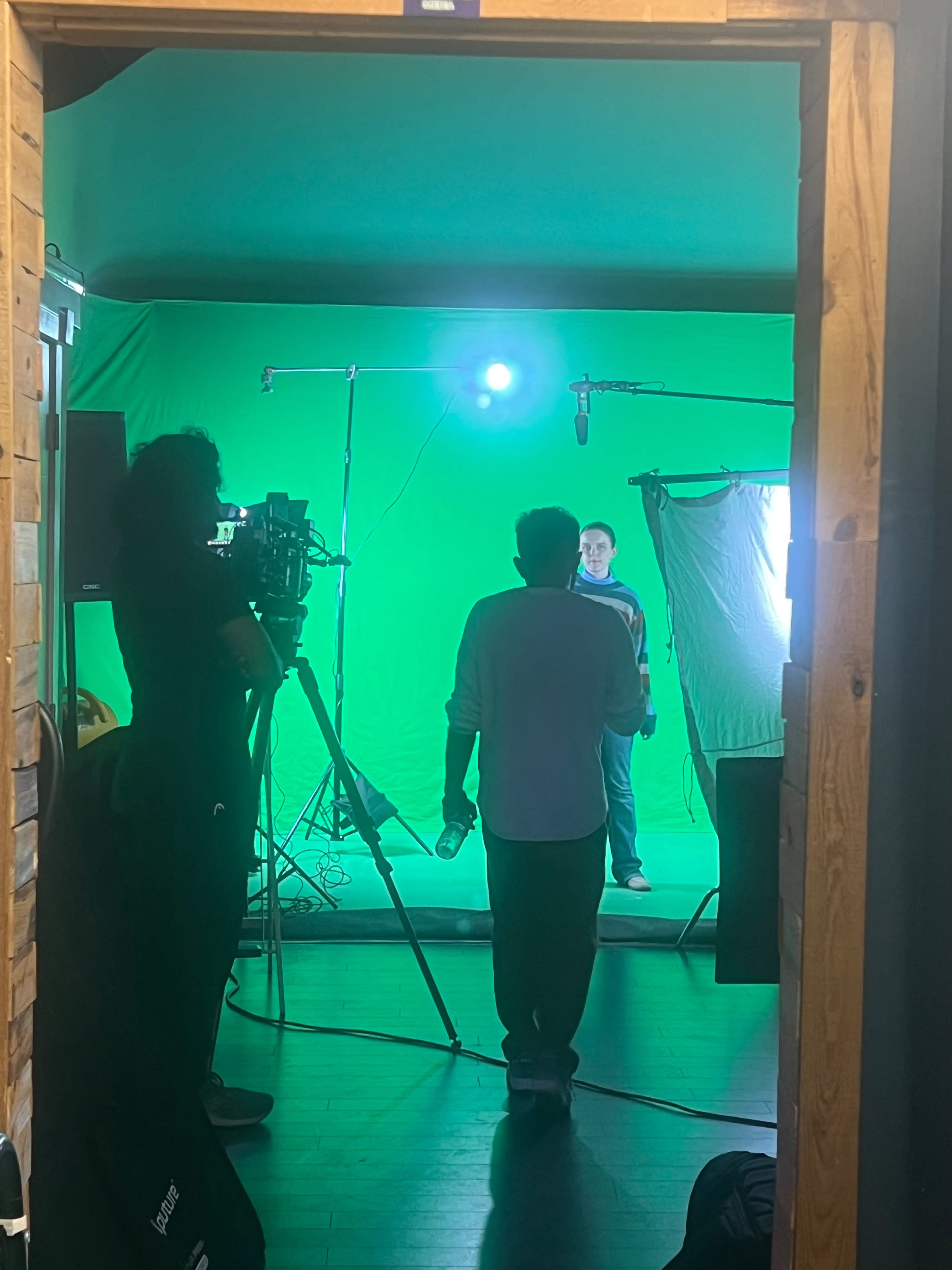 A person stands in front of a green screen as another individual operates a camera nearby.