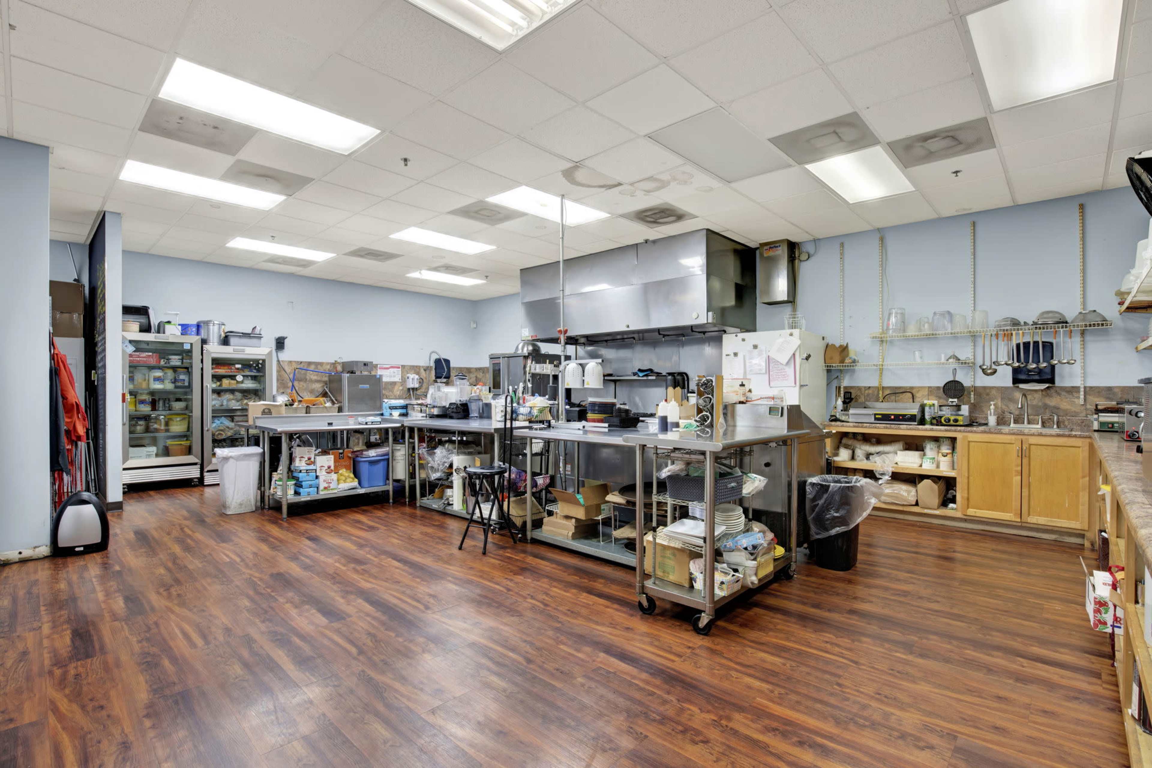 The image shows a spacious commercial kitchen with stainless steel appliances, wooden cabinets, and a variety of cooking utensils and equipment arranged on countertops.