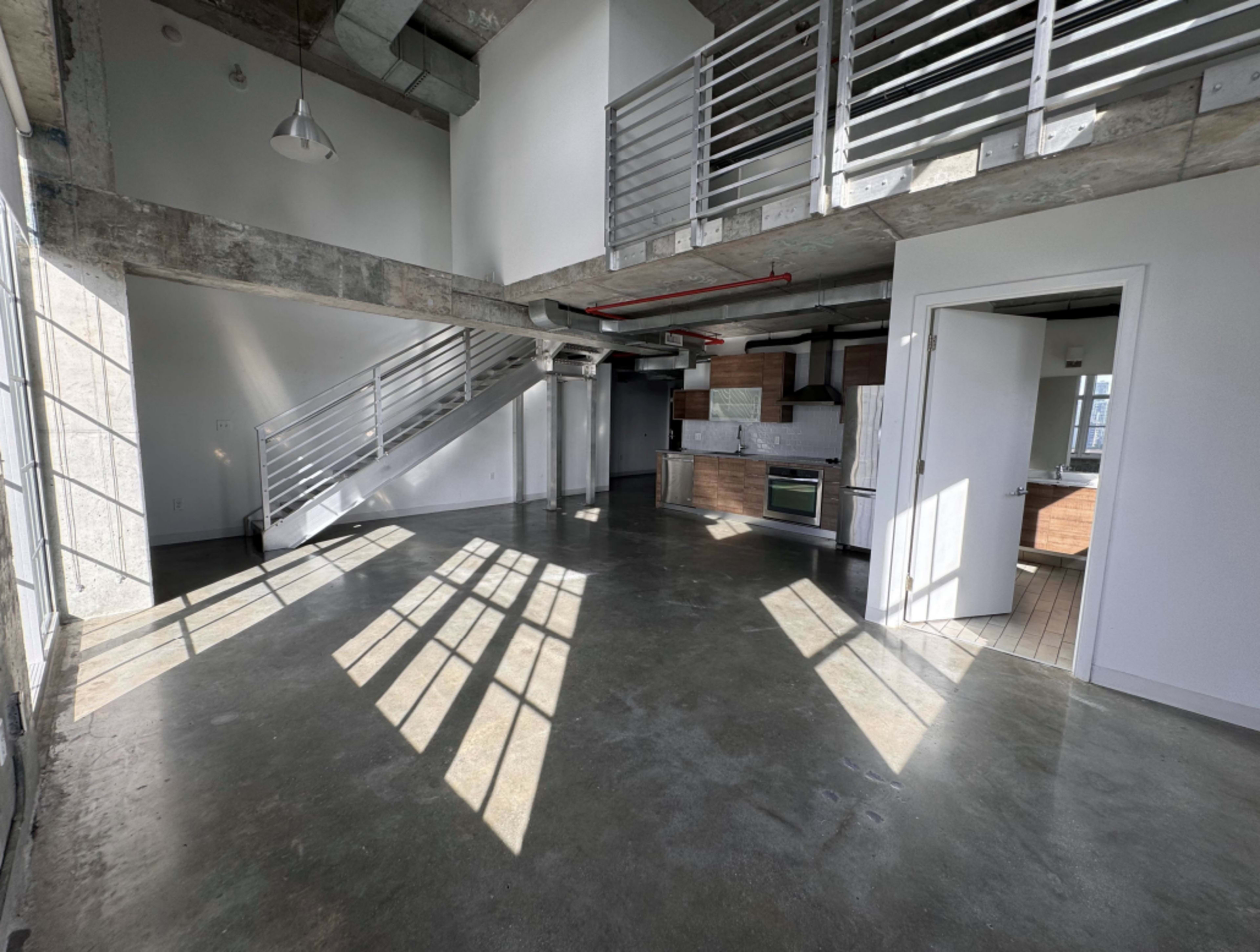 The image shows a modern, open-concept living space with industrial elements, featuring concrete floors, large windows casting shadows, and a staircase leading to an upper level.