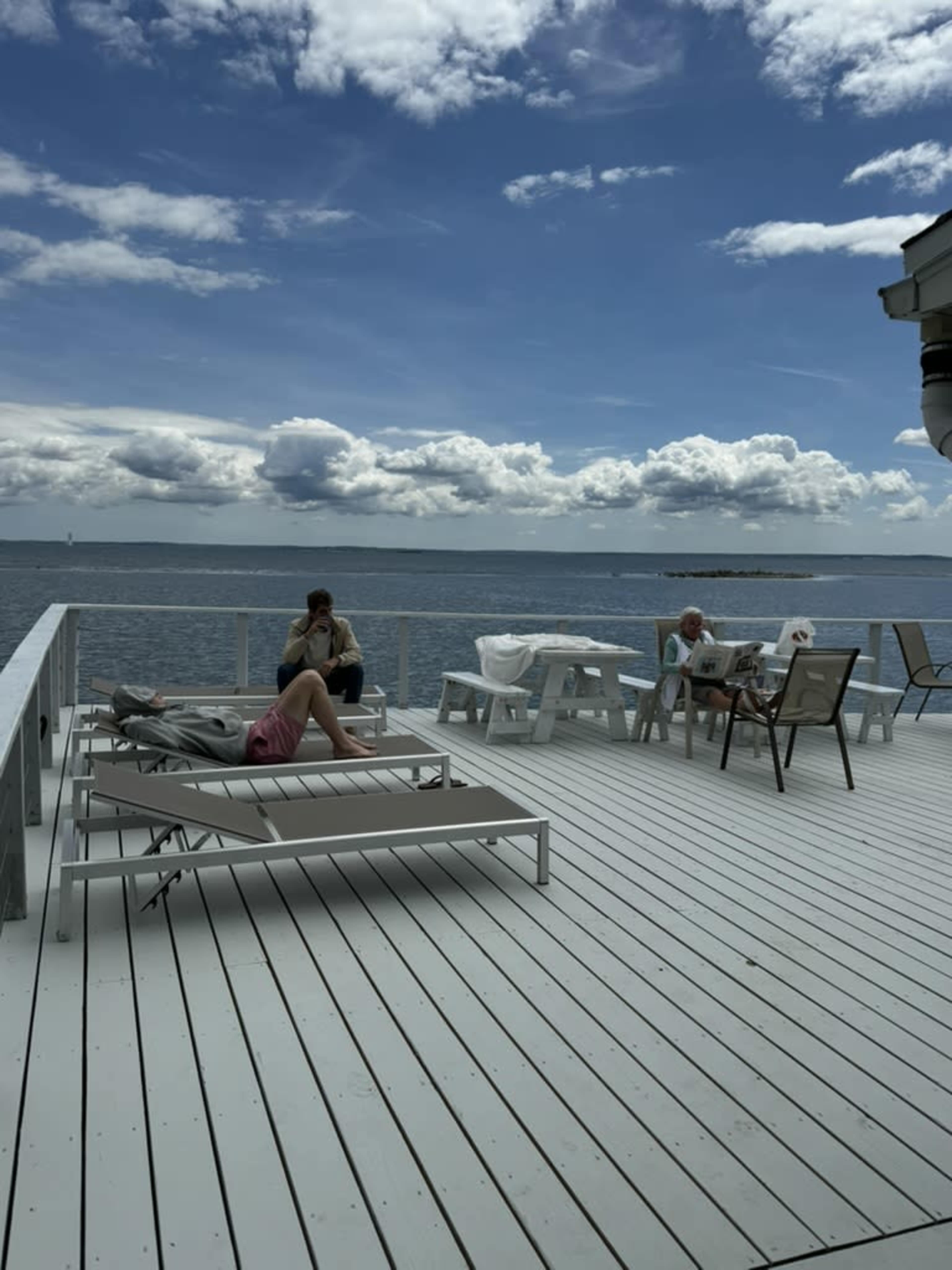 Two people relax on lounge chairs while another sits at a table on a deck overlooking the water under a cloudy sky.