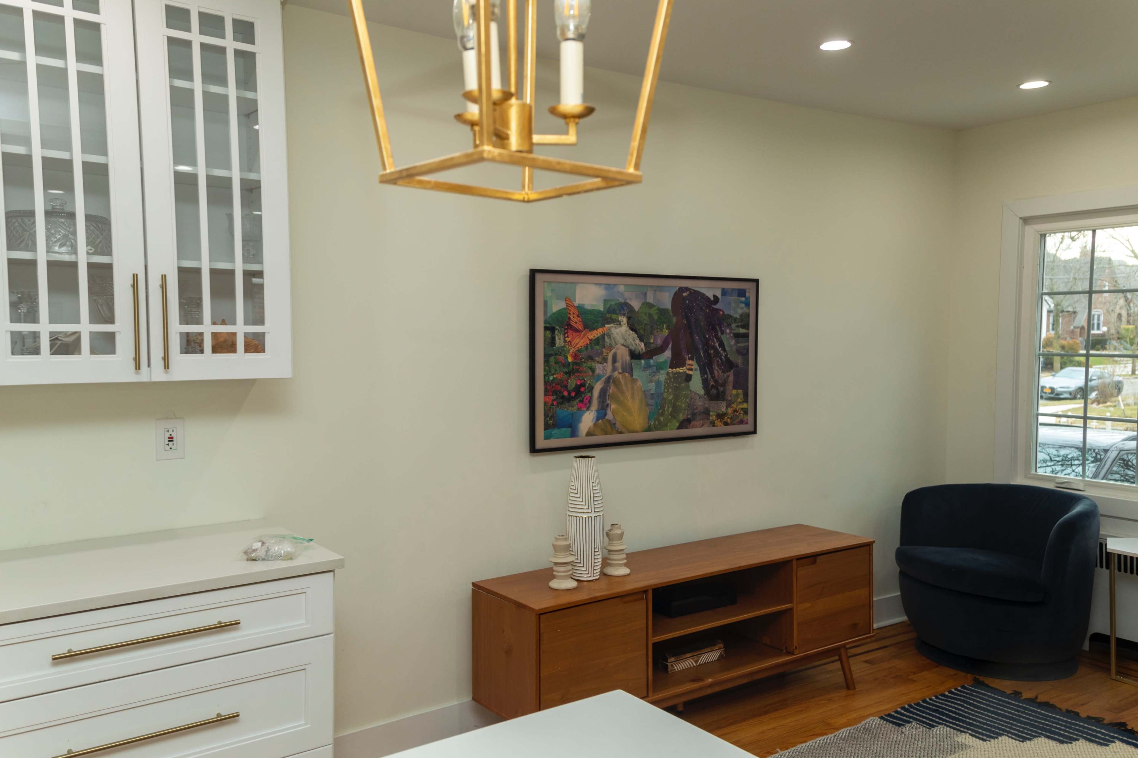 The image shows a light-colored interior featuring a wooden media console and a modern armchair, with a colorful artwork hanging on the wall.