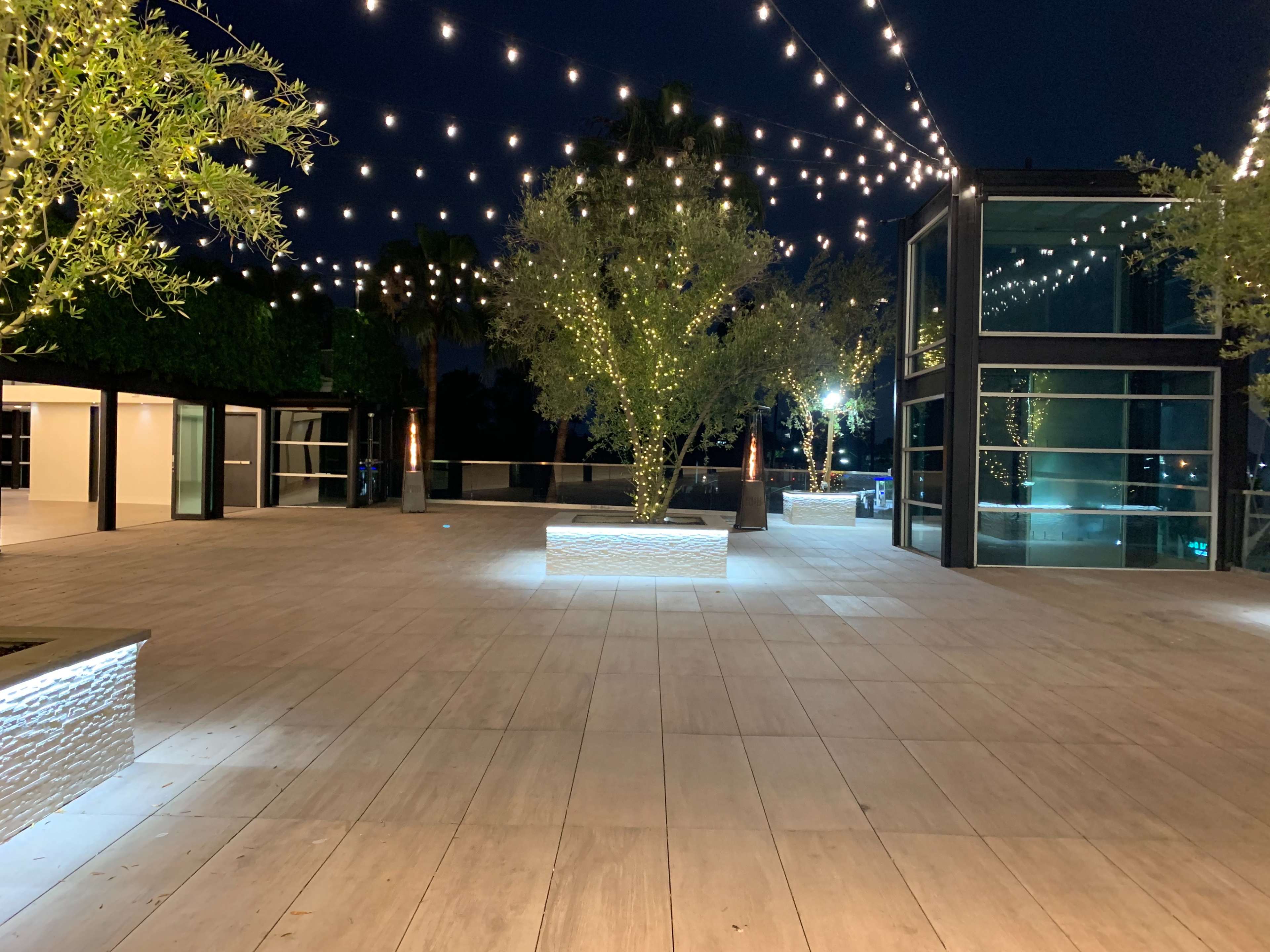 A spacious outdoor area with wooden flooring, illuminated by string lights and featuring potted trees.