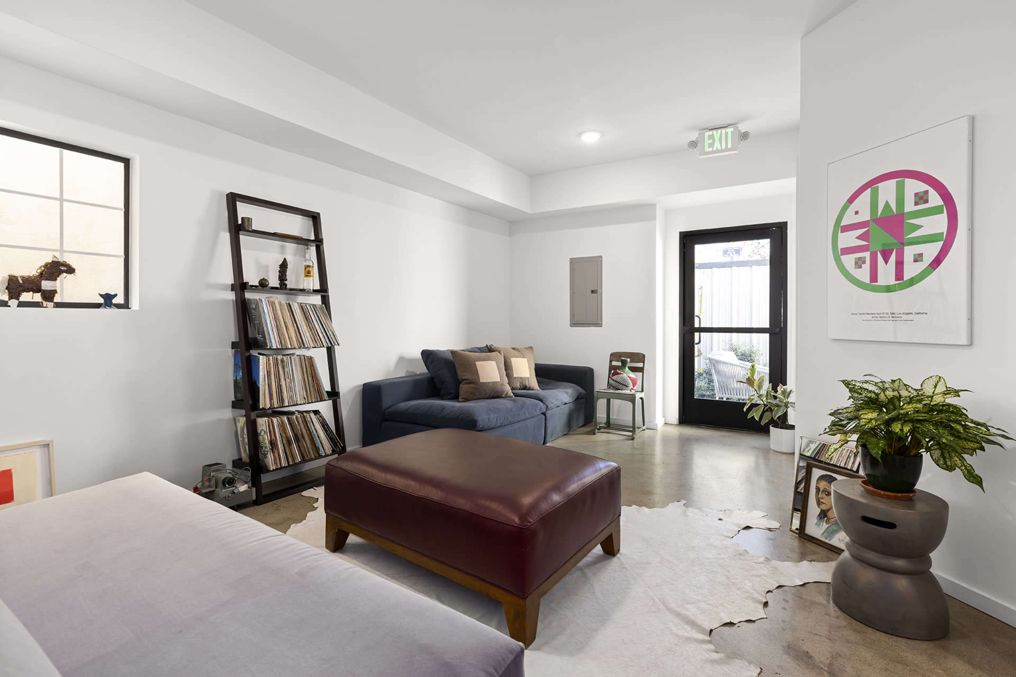 A modern living room with a blue sofa, a brown ottoman, a bookshelf filled with records, and a door leading outside.
