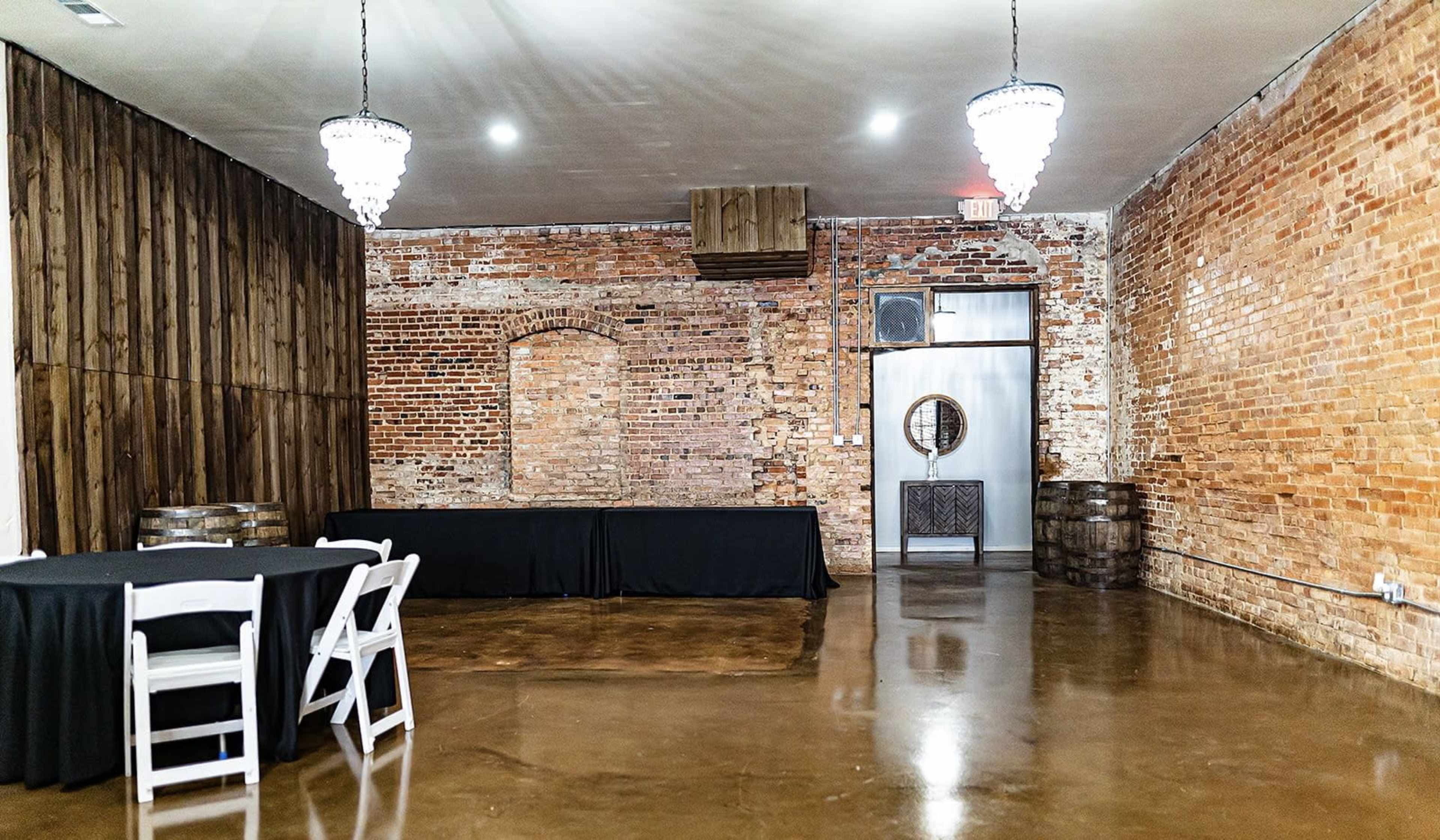 An empty room with exposed brick walls, a polished concrete floor, a table covered in a black cloth, and two chandeliers hanging from the ceiling.