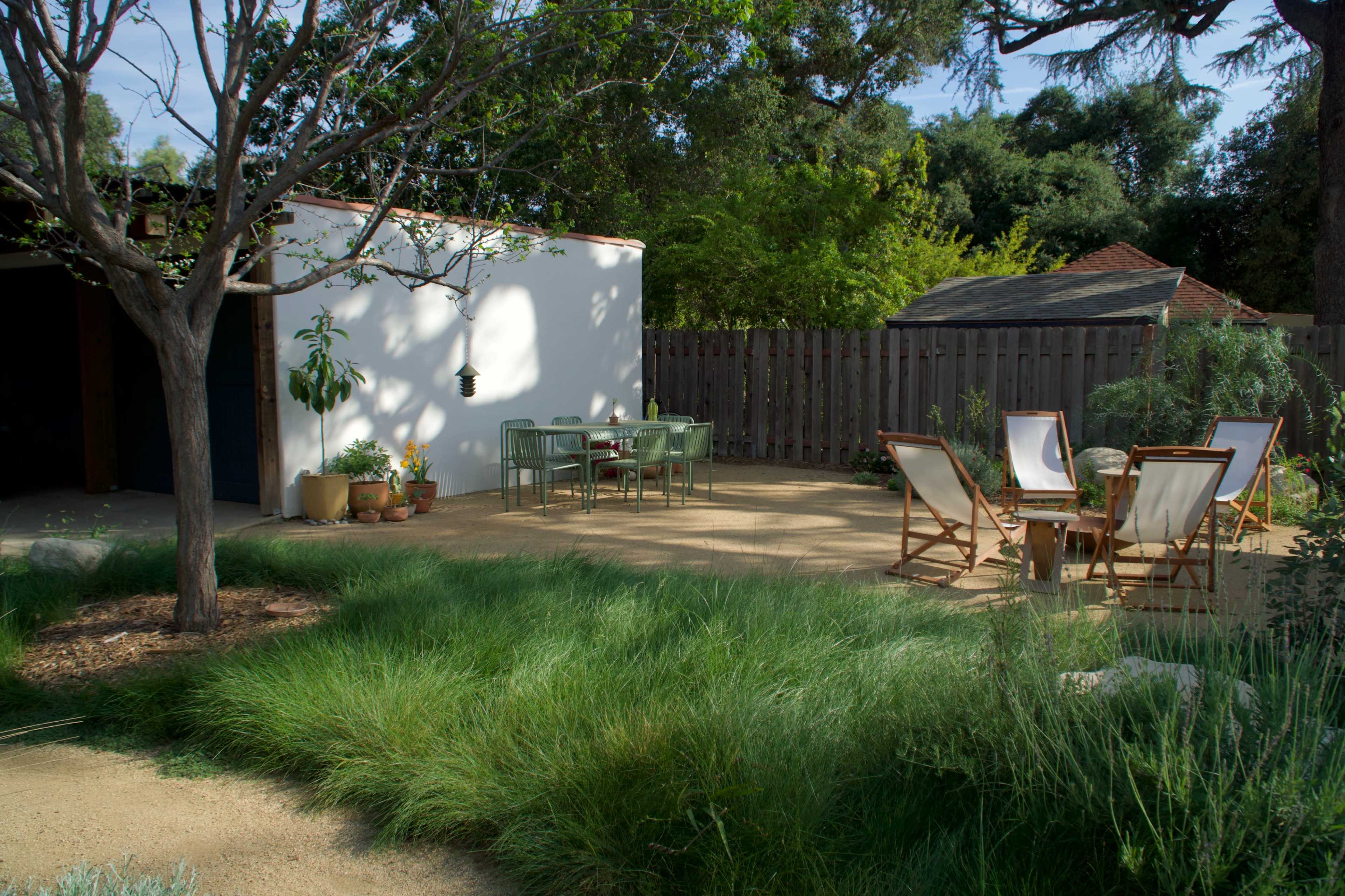 A garden patio features a dining table and chairs surrounded by tall grass and trees, with a small shed and wooden fence in the background.