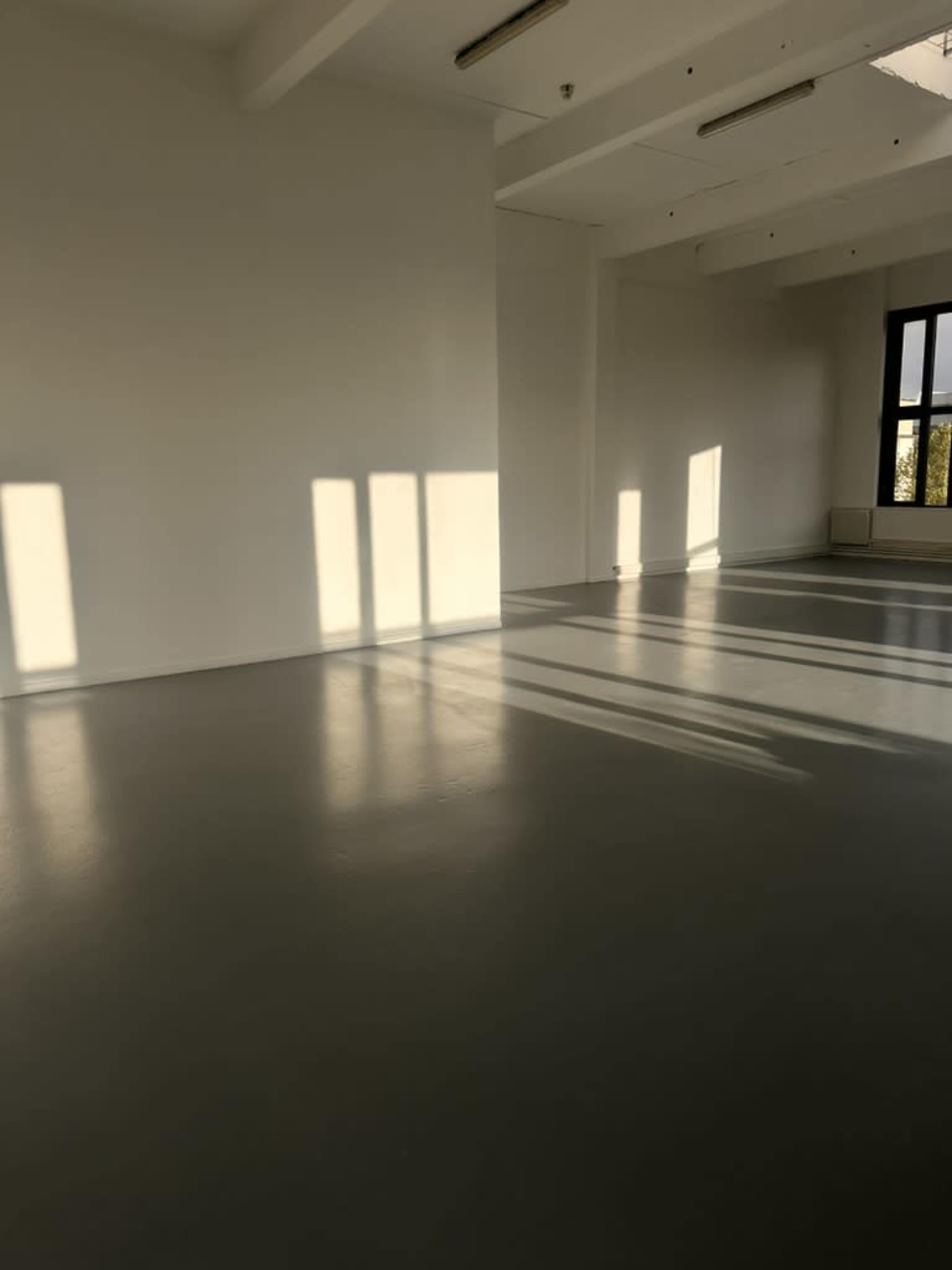 The image shows a spacious, empty room with gray flooring and sunlight casting long shadows on the walls.