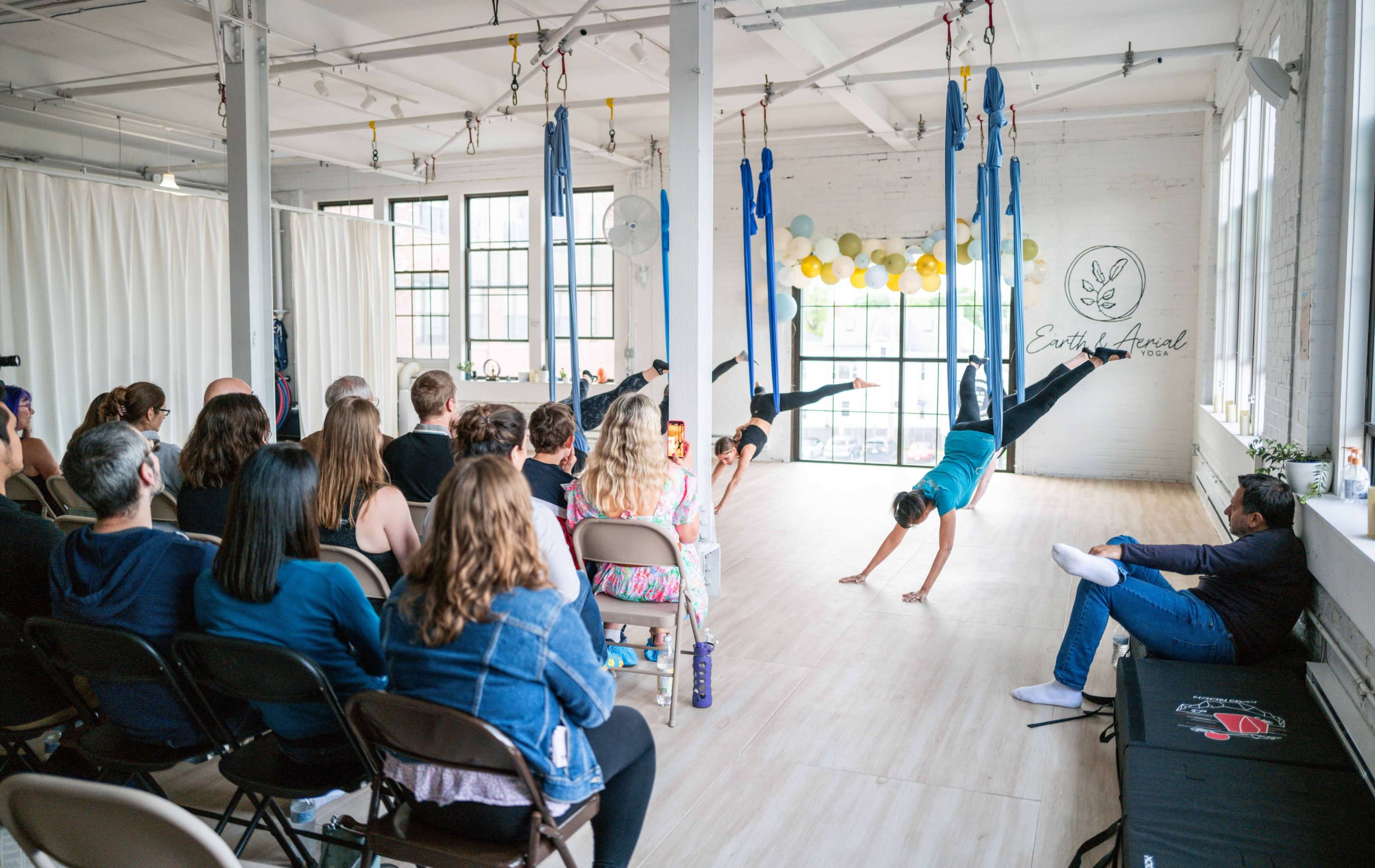 A group of people sits in chairs watching performers execute aerial routines using suspended fabric in a well-lit studio.